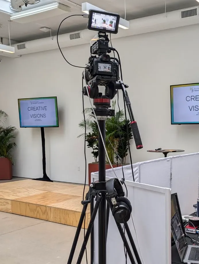 Livestream camera on a tripod in front of a small stage with two TV screens