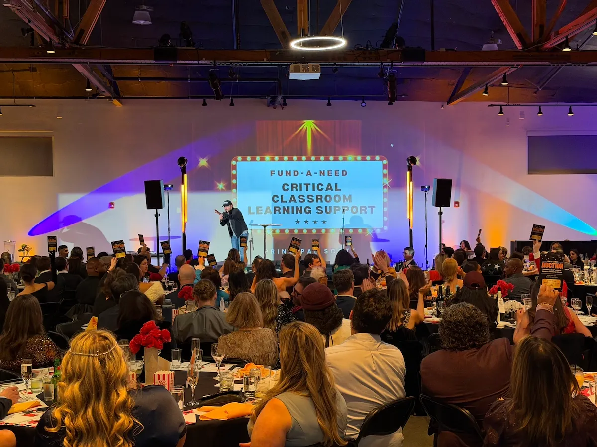 Conference audience at round tables facing a stage with two projection screens and uplighting