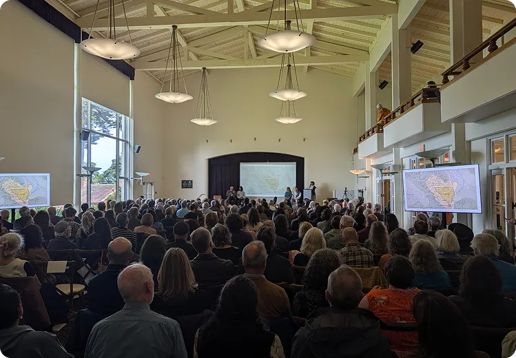 Large audience in a hall watching speakers on stage and side screens