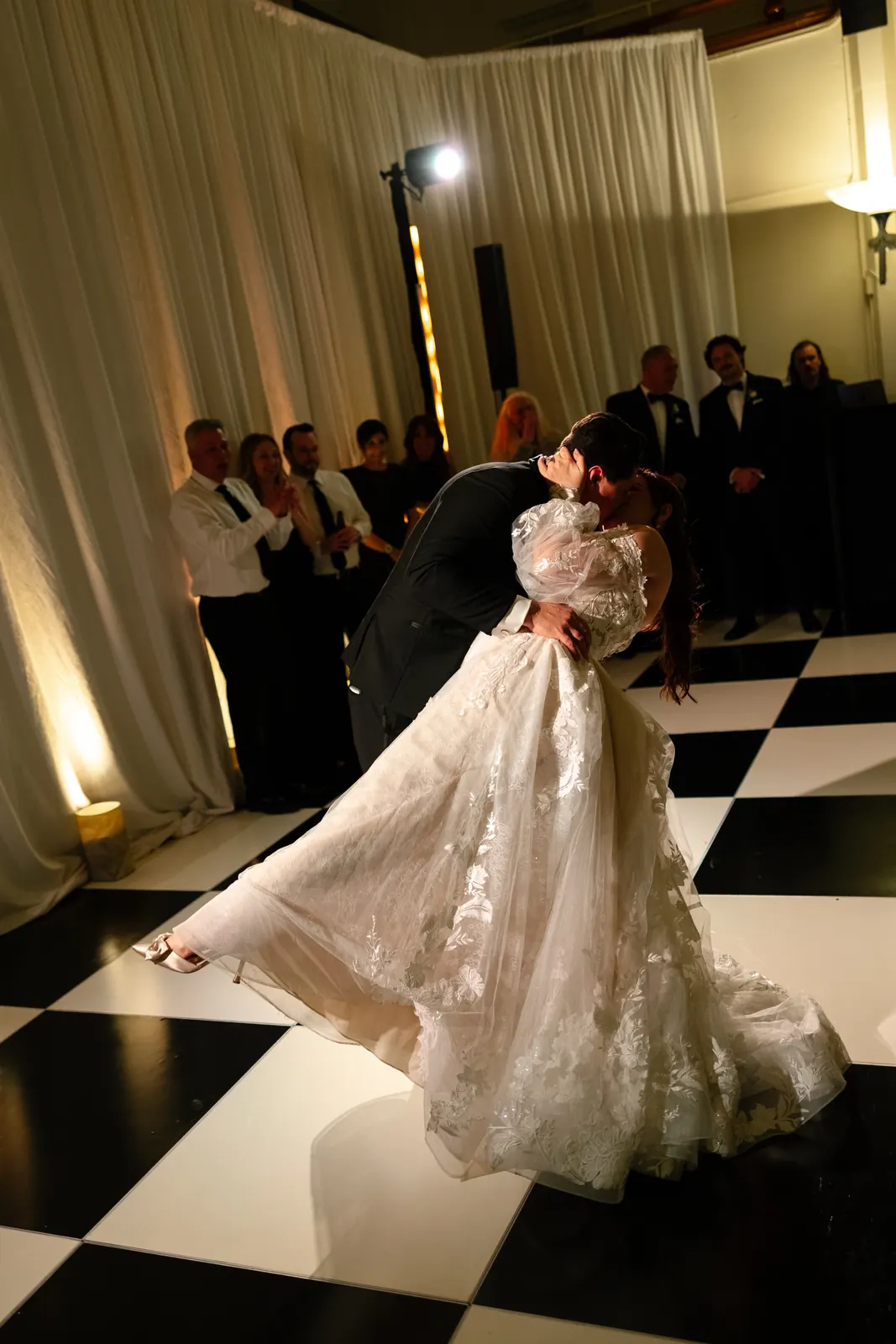 Couple's first dance on a black-and-white checkered floor, with warm uplighting on white drapes