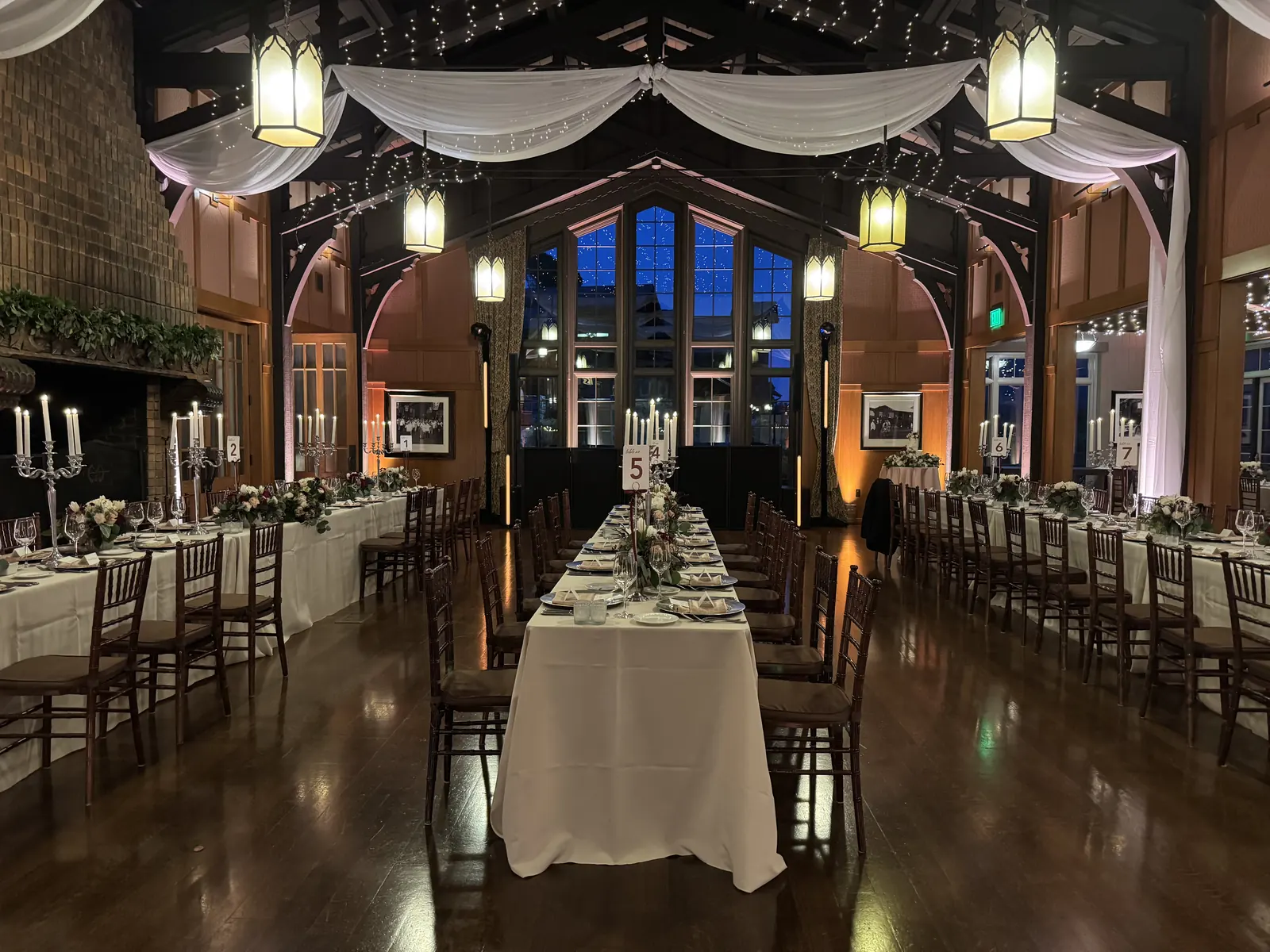 Long reception tables in a wood-beamed lodge with draped fabric overhead and warm lantern light