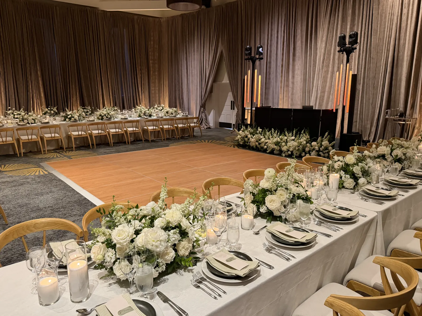 Wedding reception set with a wood dance floor, white floral tables and speakers on stands against tan drapes