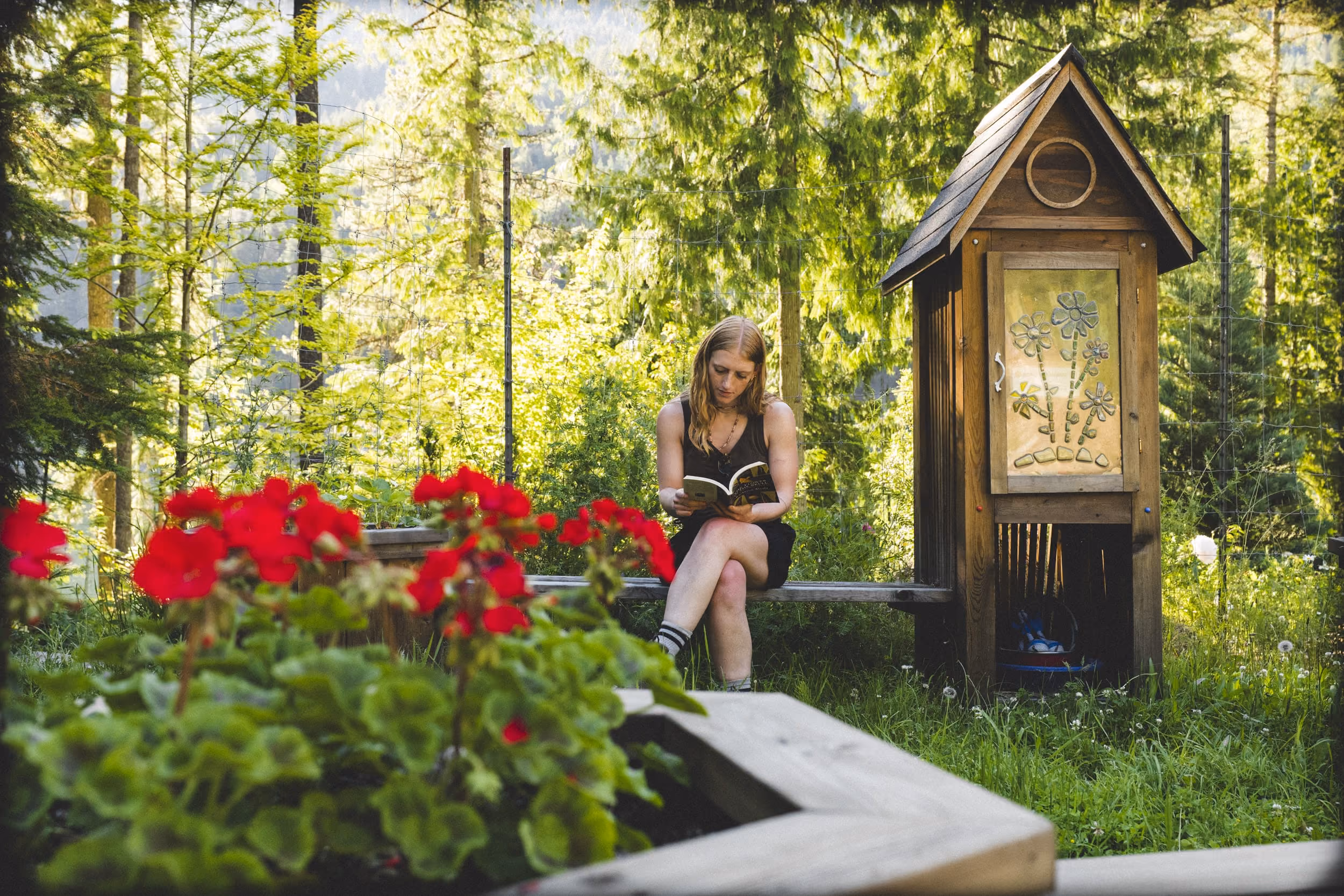 Woman reading a book on a bench in a garden with red flowers and trees around.