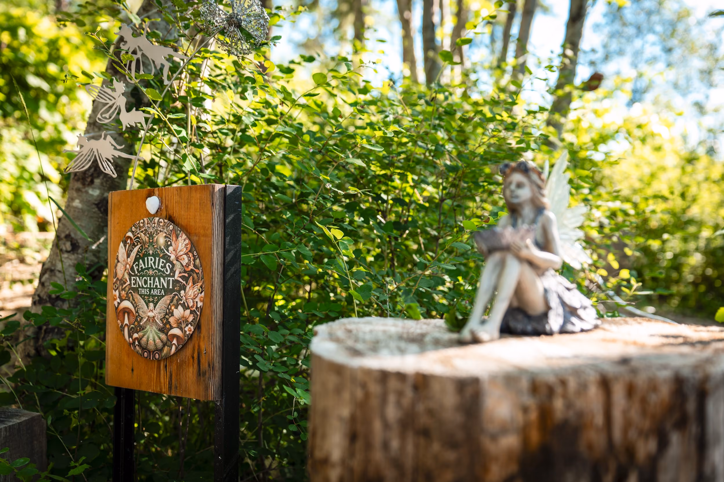 Wooden sign reading 'Fairies Enchant This Area' beside a small fairy statue on a tree stump outdoors.