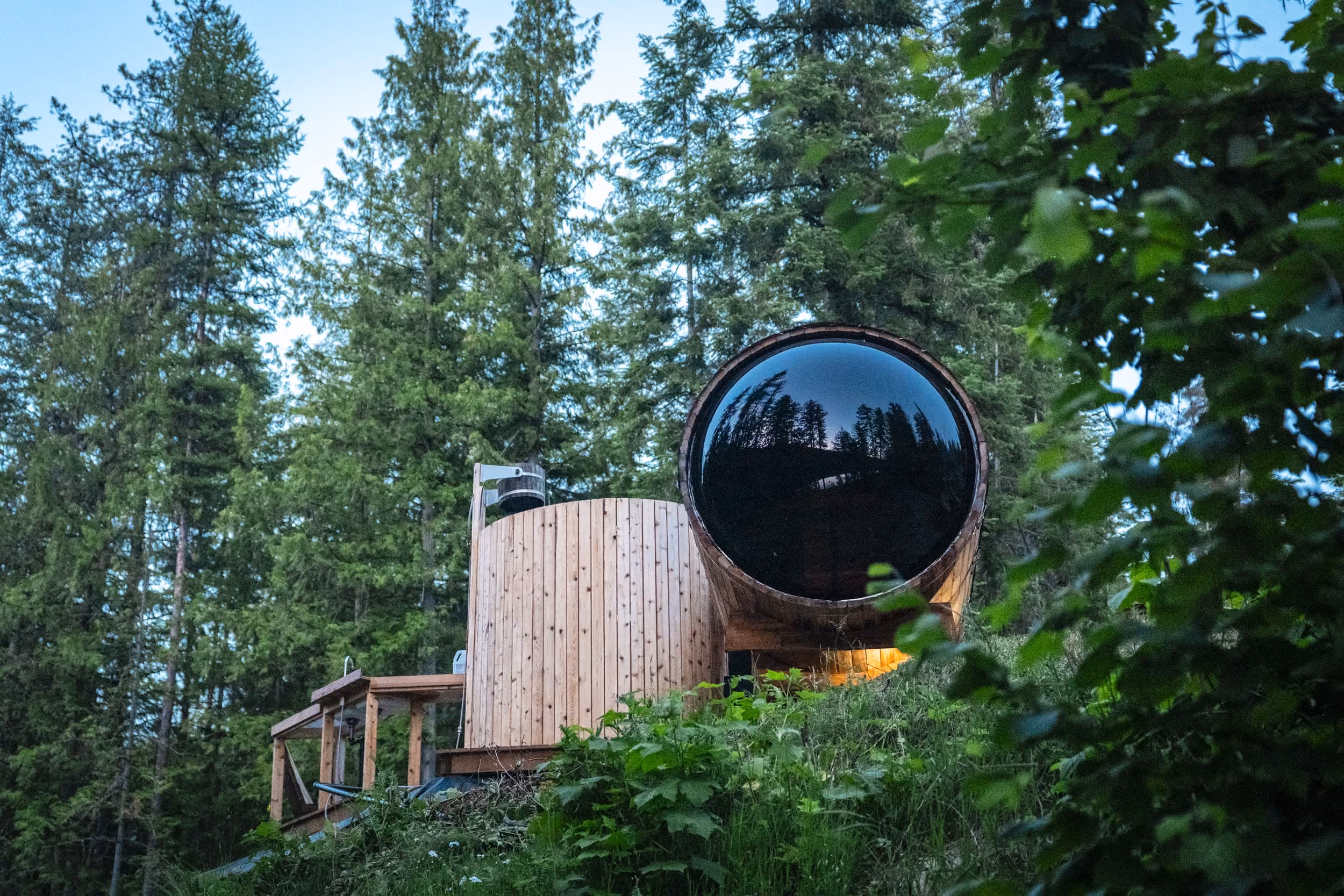 Wooden sauna with large circular glass front reflecting trees, surrounded by forest greenery on a hillside.