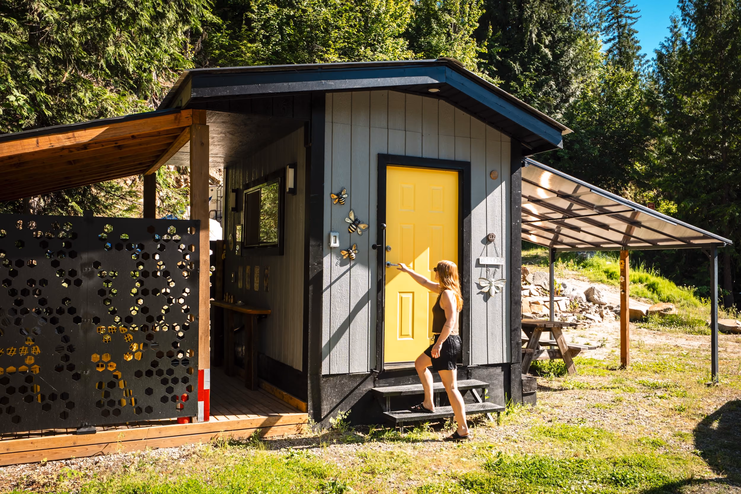 Woman wearing black clothes reaching to open yellow door of a small gray cabin with porch and carport.