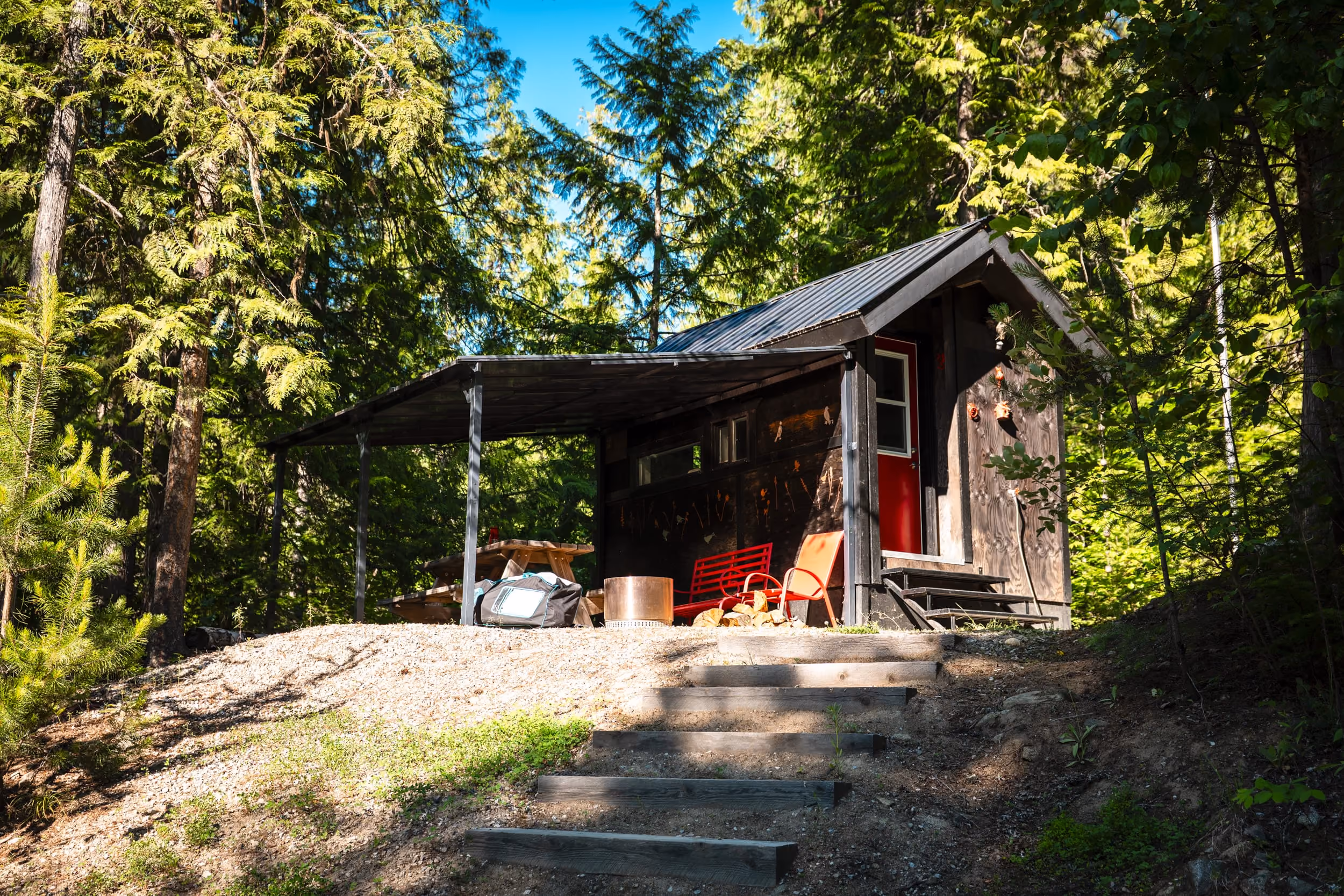 Small dark wooden cabin with red door and chairs under a metal roof in a dense forest.