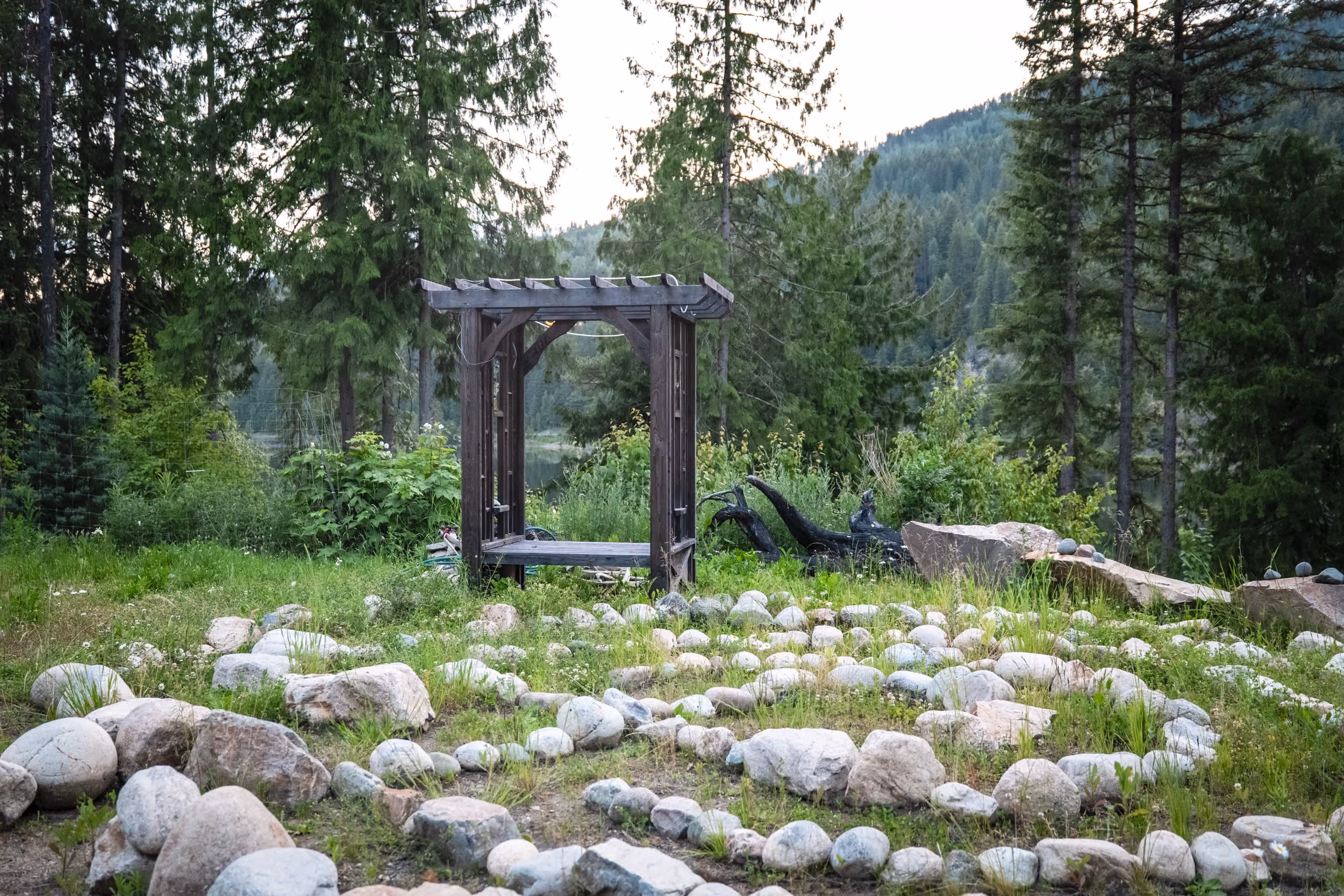 Stone labyrinth path on grass with wooden pergola and forested mountain background.