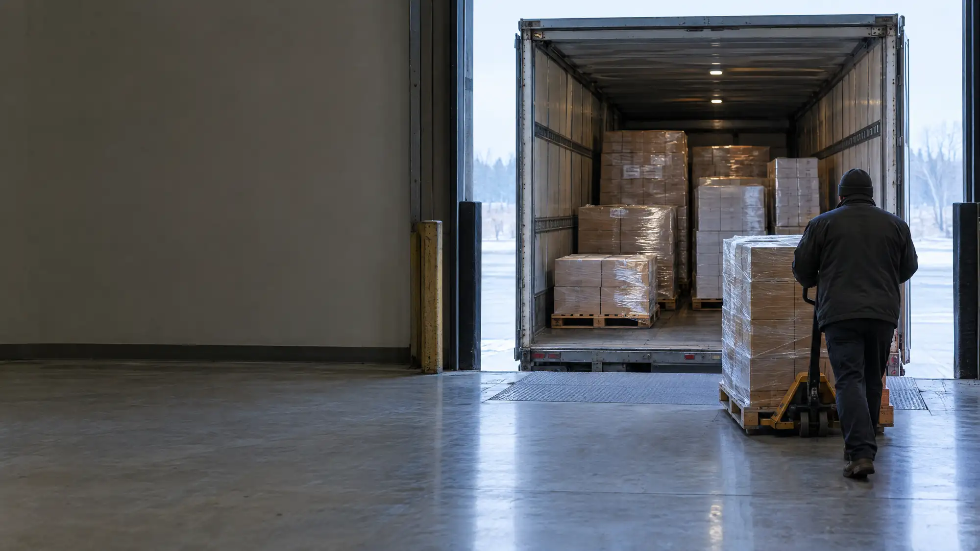 Warehouse worker pushing a shrink-wrapped pallet of cartons toward a partly loaded LTL trailer at an open dock door
