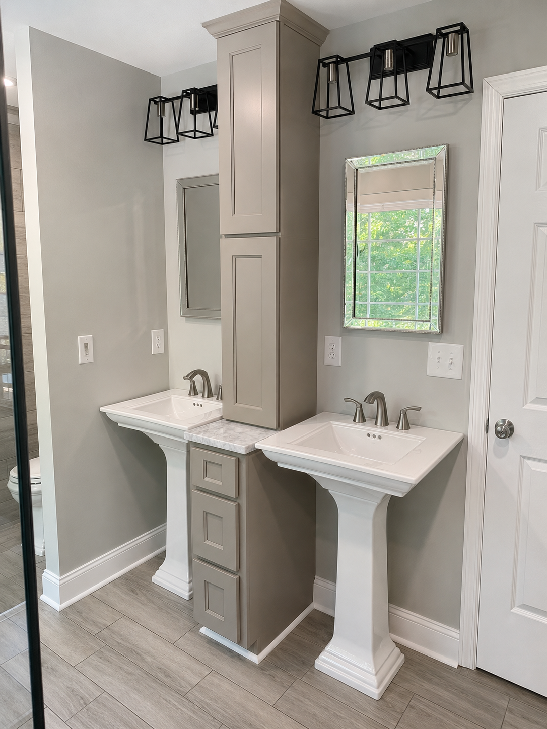 Two white pedestal sinks separated by a gray floor-to-ceiling storage cabinet with a stone countertop and framed mirrors in Davidson, NC.