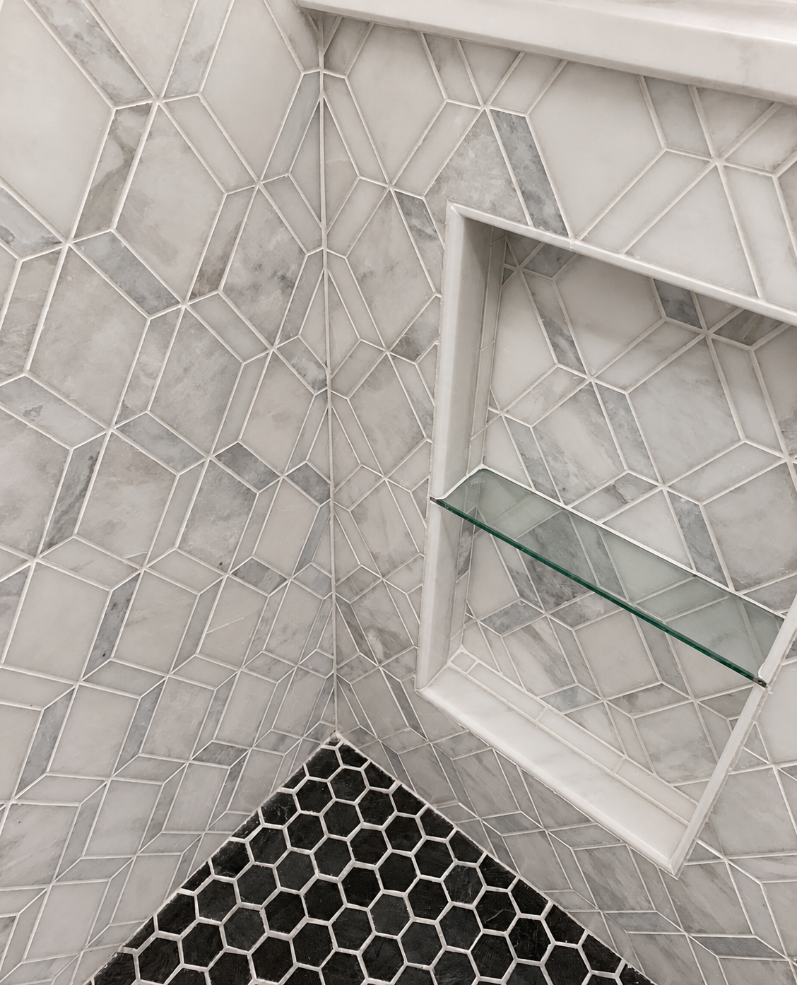 Close-up of a geometric marble mosaic shower corner with a recessed niche, glass shelf, white trim, and black slate hexagon floor tile in Fort Mill, SC.