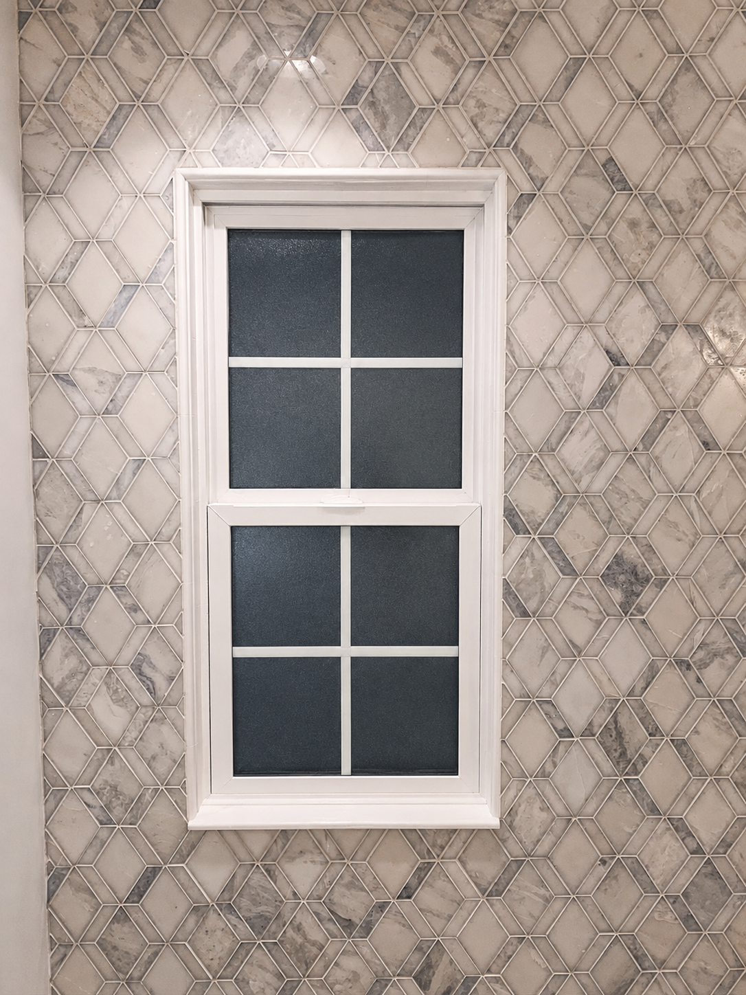 Bathroom window surrounded by white and gray geometric mosaic tile with carefully fitted cuts around the trim in Fort Mill, SC.
