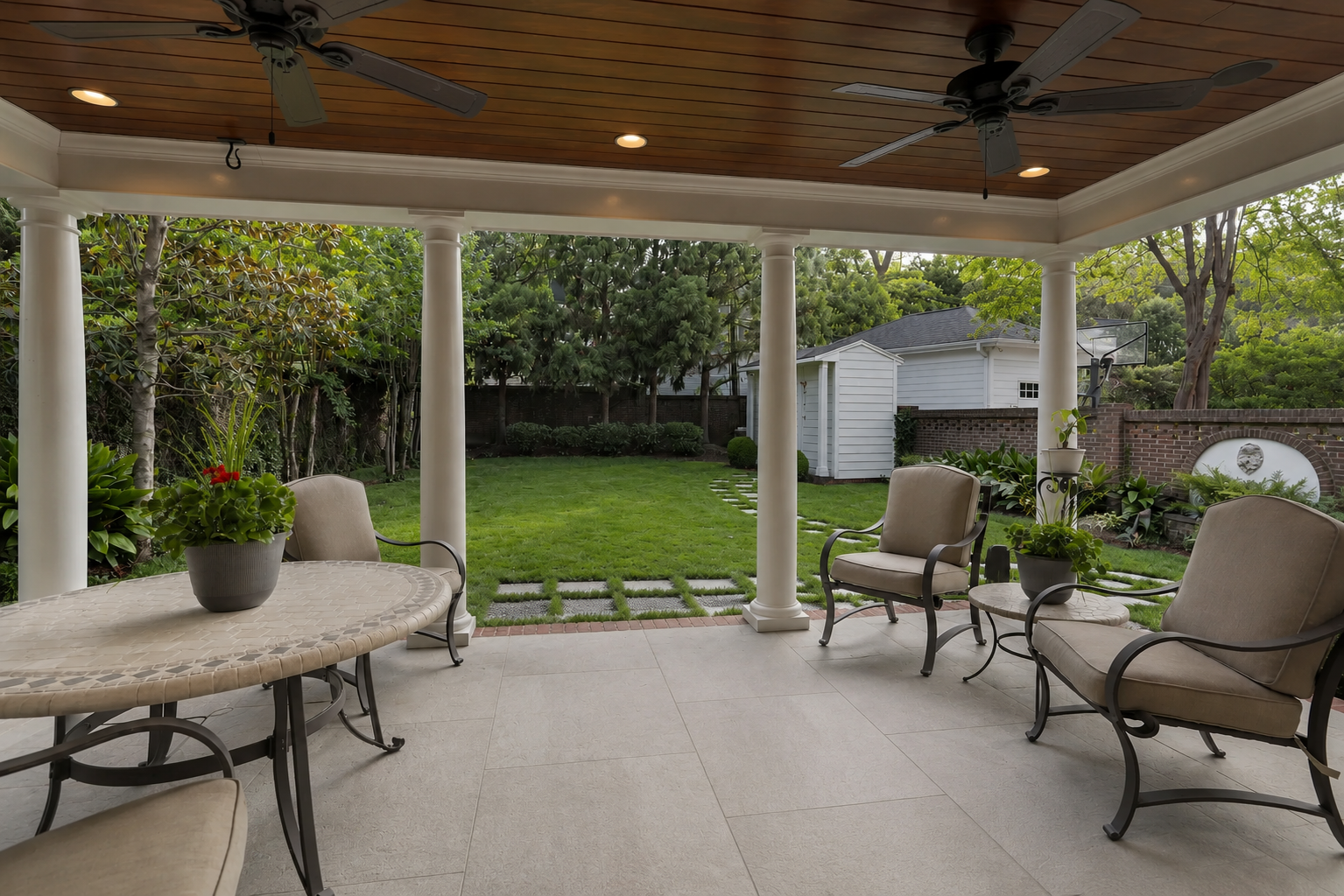 Covered outdoor patio with large-format light neutral tile flooring, white columns, a wood-plank ceiling, and garden views in Mooresville, NC.