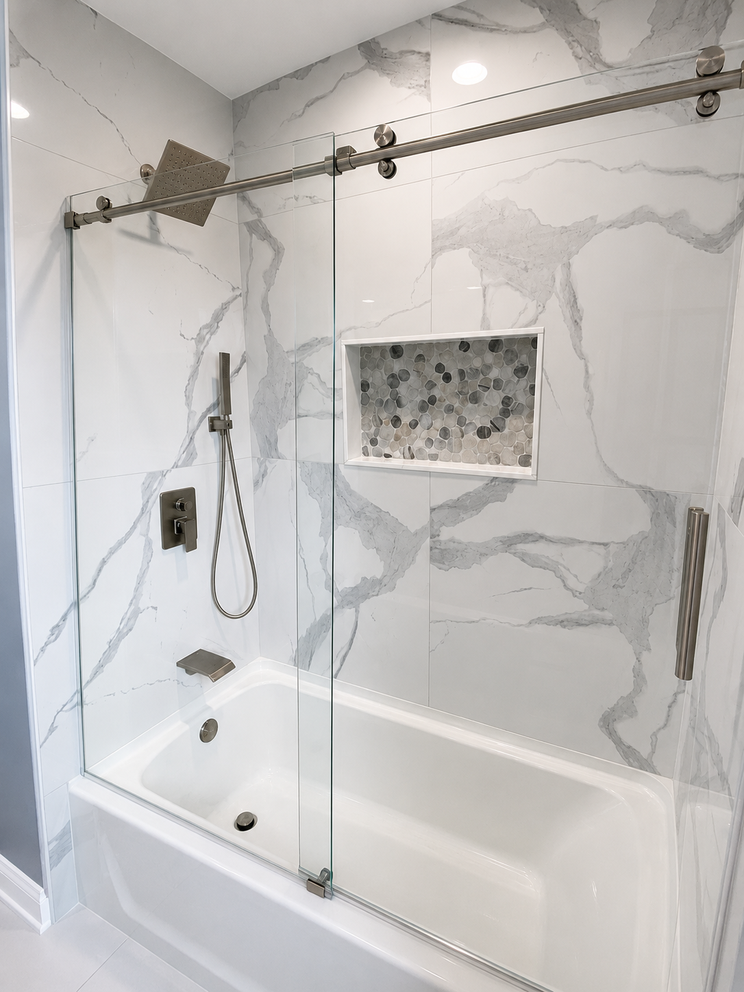 Bathtub and shower surround with large-format white marble-look tile, a pebble mosaic niche, rainfall showerhead, and clear sliding glass enclosure in Stallings, NC.