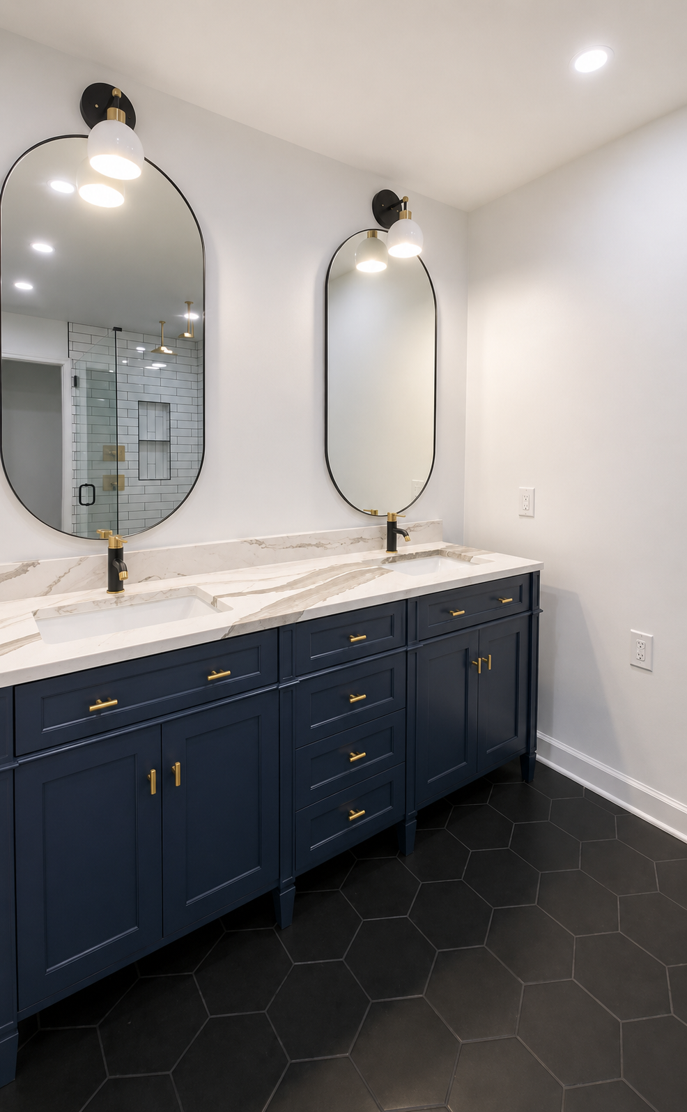 Double navy bathroom vanity with white marble-look countertop, arched mirrors, brushed brass fixtures, and large black hexagon floor tile in Fort Mill, SC