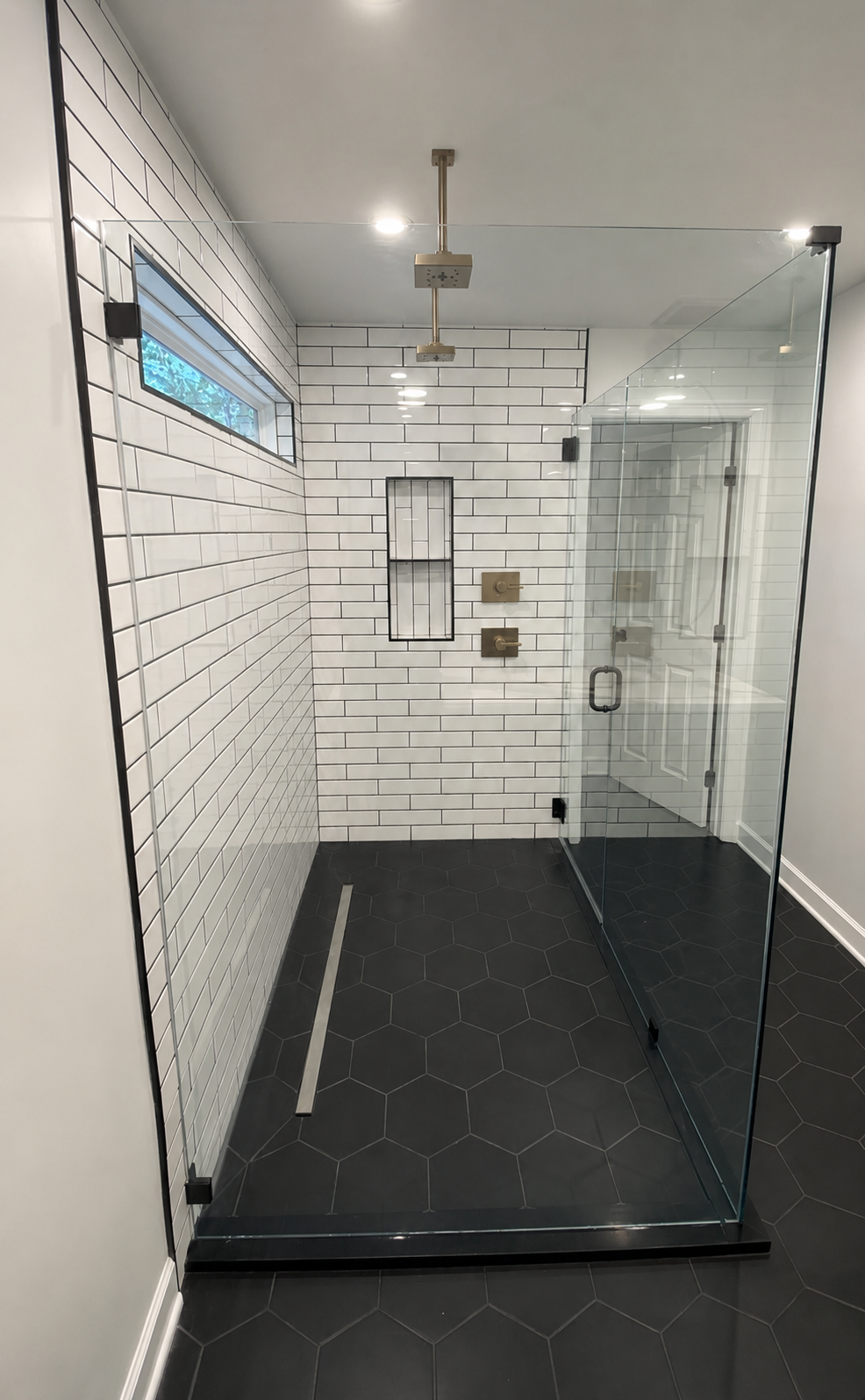 Corner view of a frameless glass shower with full-height white subway tile, black grout, black hexagon flooring, and brass ceiling-mounted shower fixtures in. Fort Mill, SC