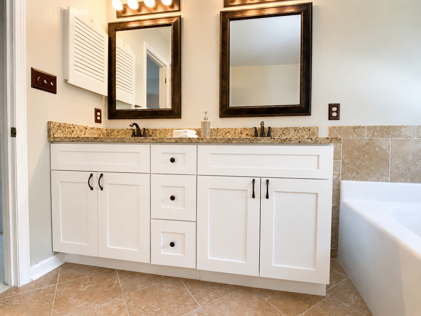White double bathroom vanity with granite countertop, dark framed mirrors, bronze fixtures, beige floor tile, and tiled bathtub surround in Concord, NC.