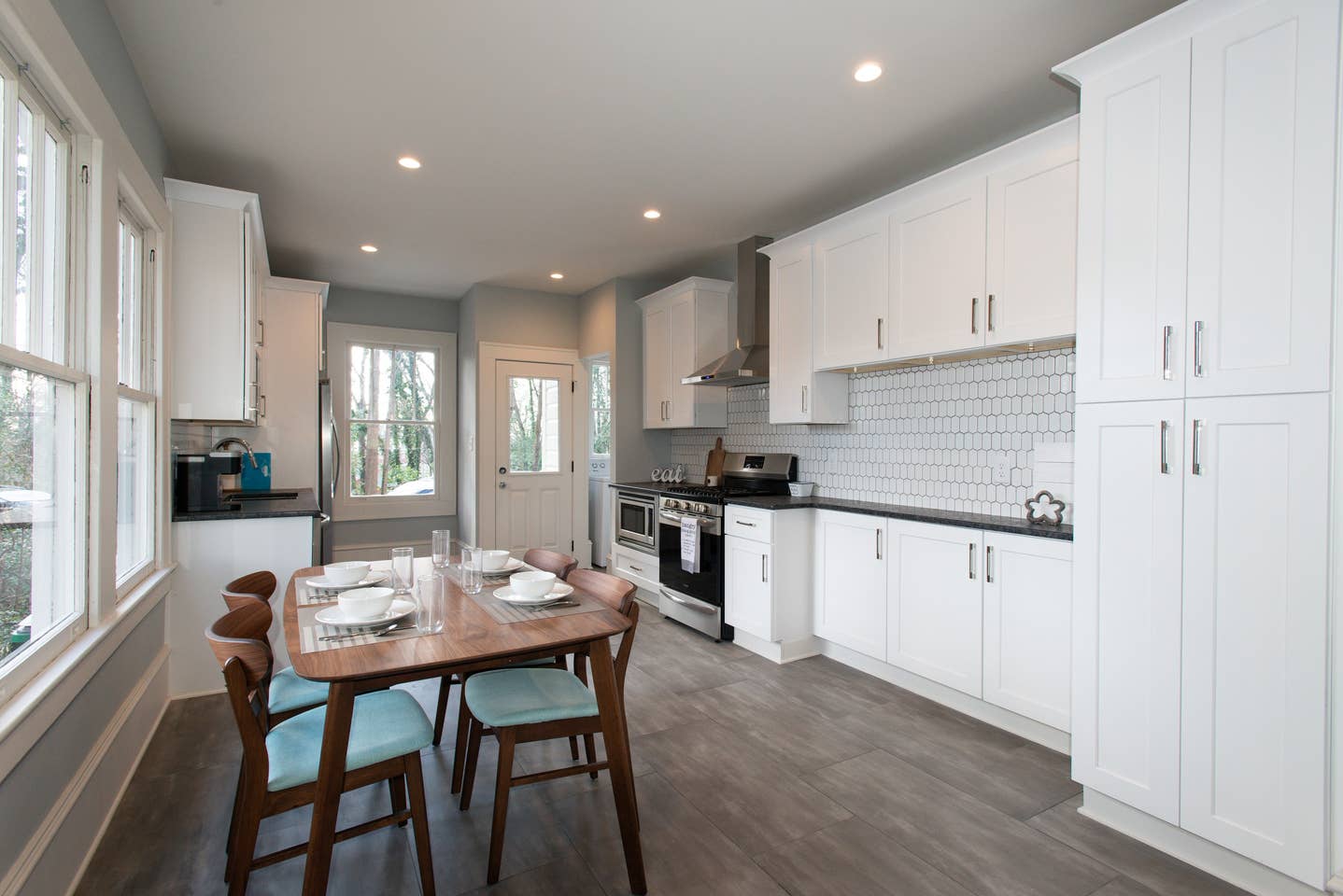 Bright white kitchen with shaker cabinets, dark countertops, stainless appliances, elongated white hexagon backsplash tile, dining table, and gray plank flooring in Charlotte, NC located in the Wilmore/Southend district