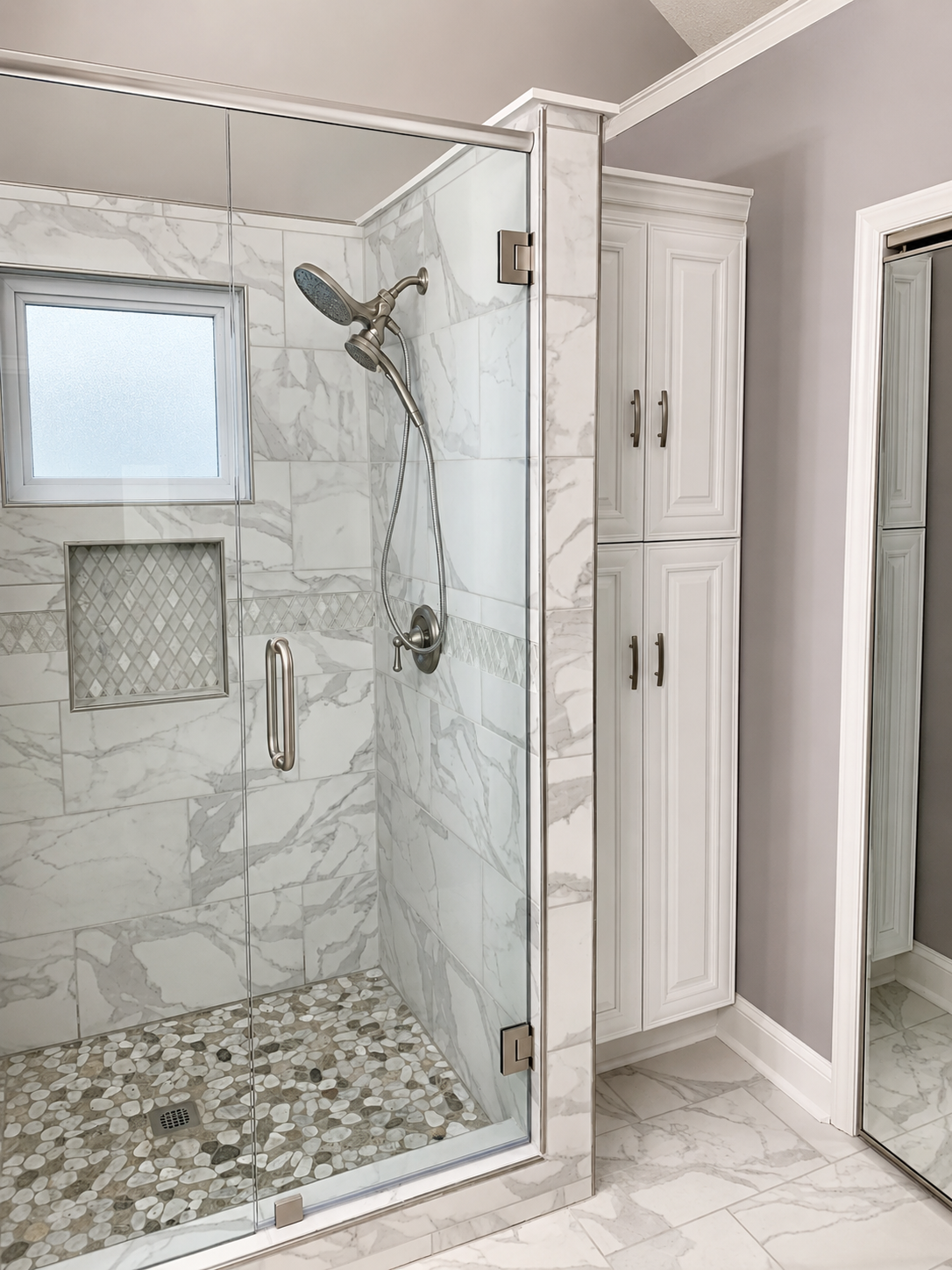 Alternate view of a frameless marble-look tiled shower with pebble mosaic floor, decorative niche, brushed metal fixtures, and matching bathroom floor tile in Concord, NC