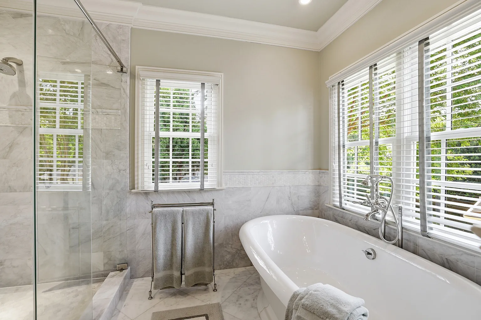 Bright primary bathroom with a glass shower, white-and-gray marble surfaces, decorative mosaic trim, freestanding bathtub, chrome fixtures, and large windows in Davidson, NC.