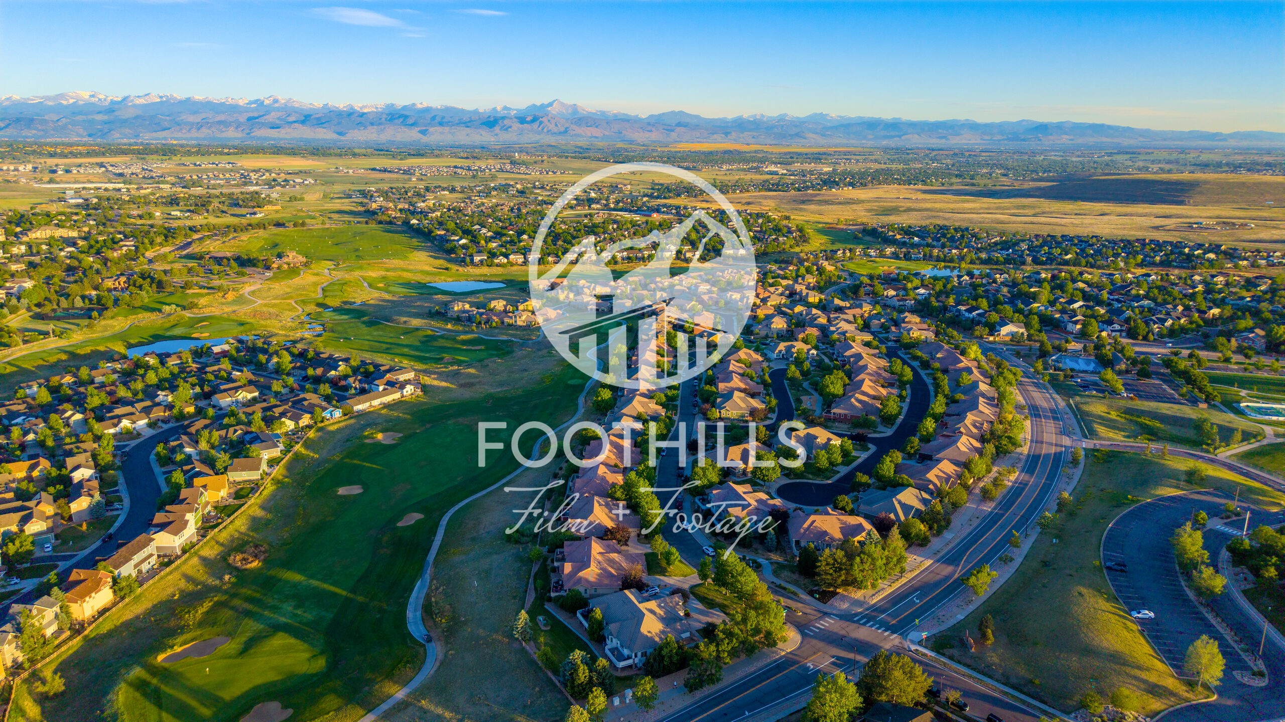 Golden-hour aerial of Vista Ridge in Erie, Colorado, with curved streets wrapping around Colorado National Golf Club and its ponds, and the snow-capped Front Range across the horizon