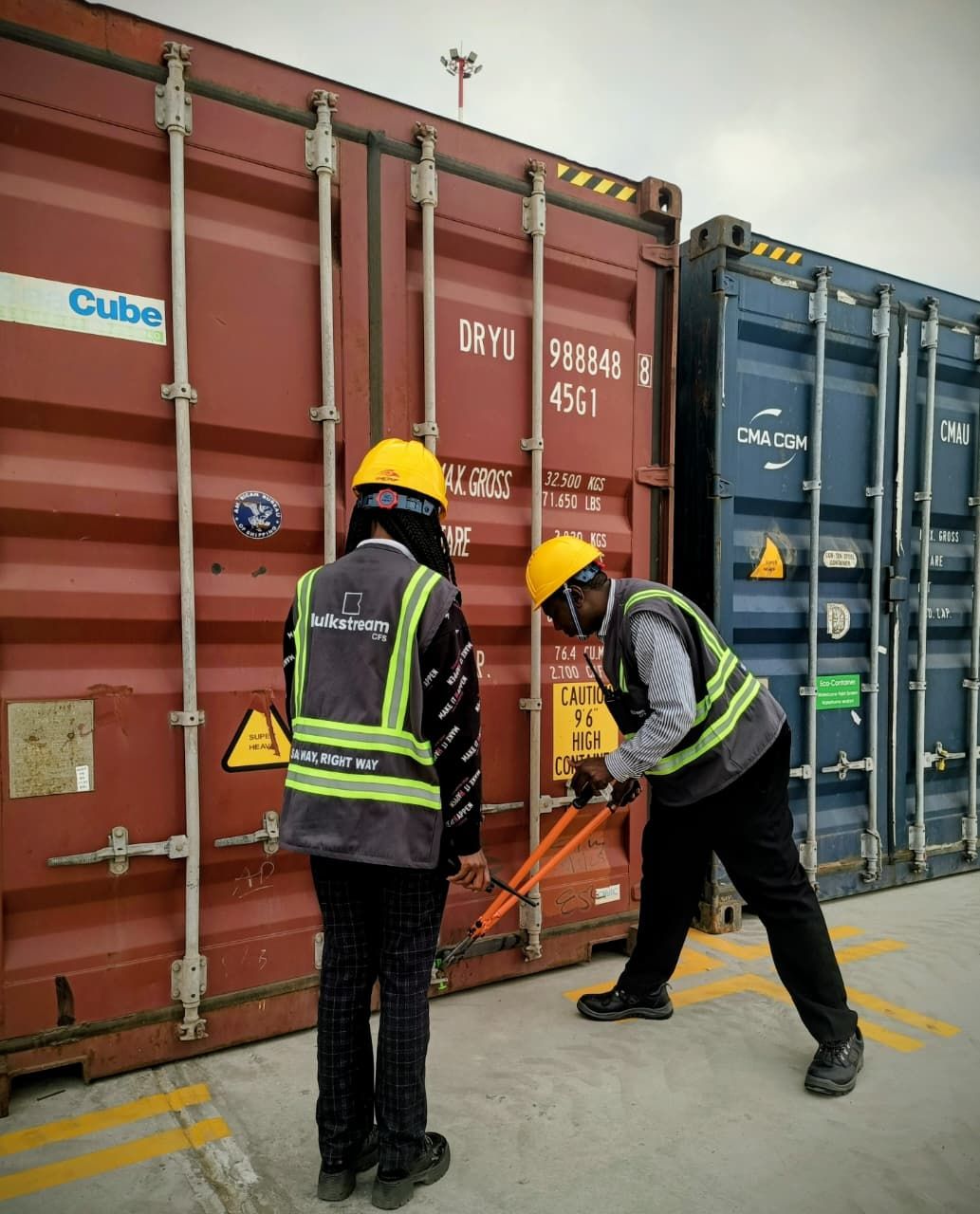 Two staff in hard hats with bolt cutters at a container door in the yard