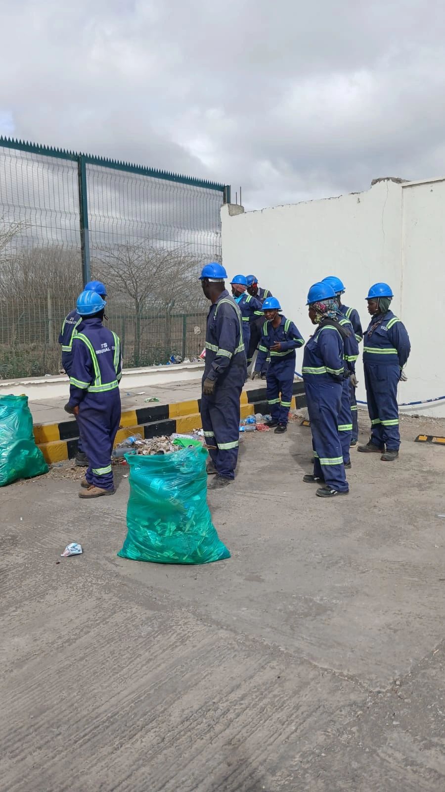 Team members picking up litter beside the perimeter fence, with a filled waste bag in front