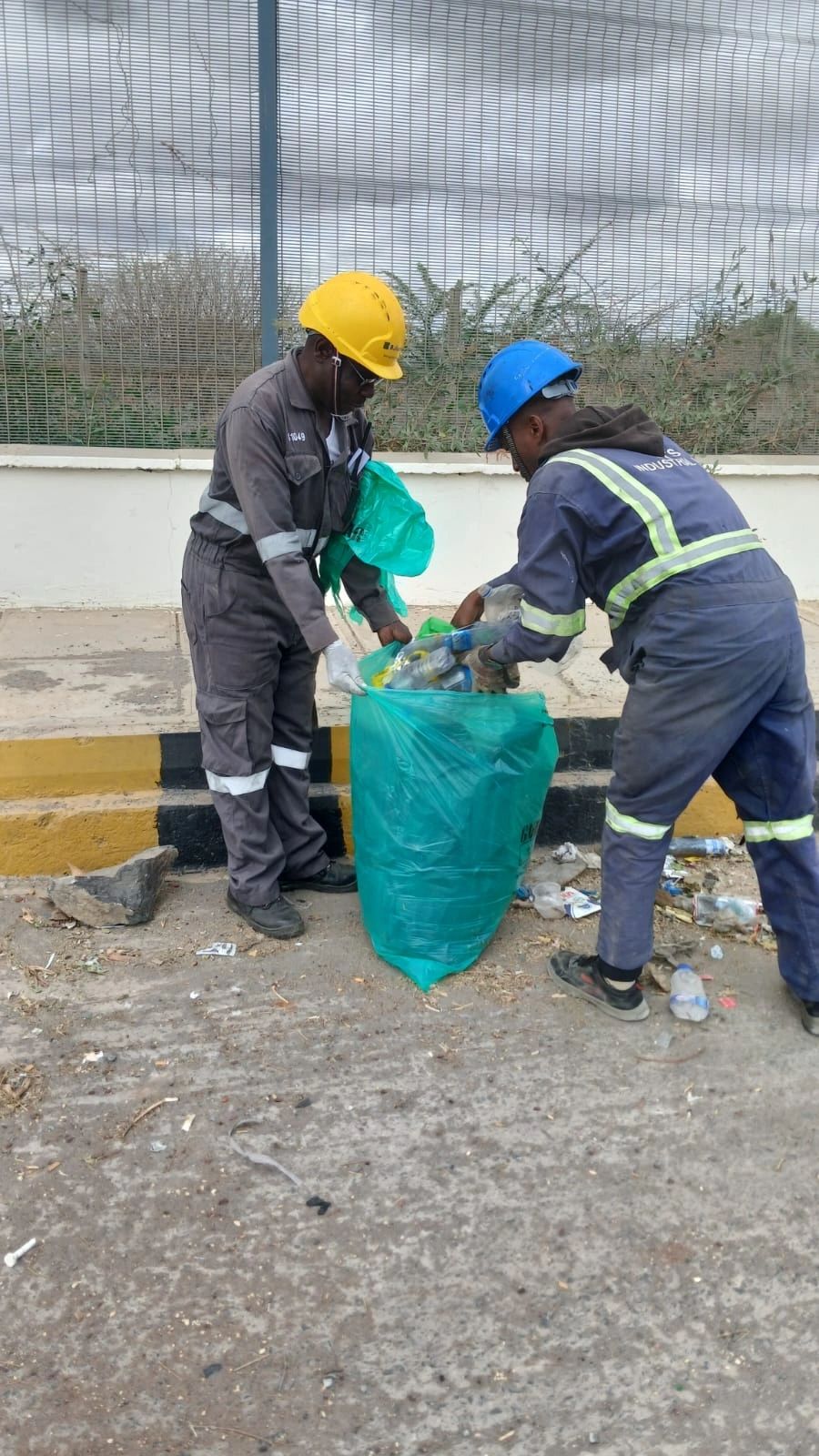 Two team members in hard hats and gloves collecting plastic litter into a waste bag