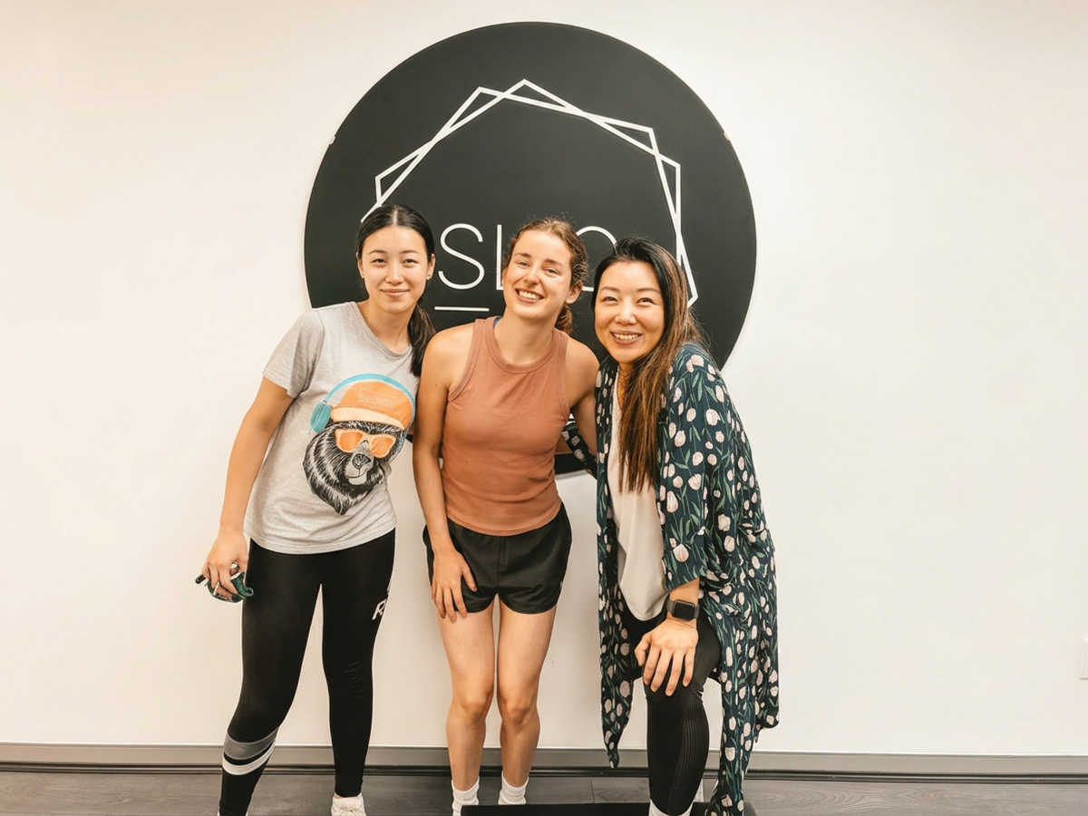 Three smiling women standing together in front of the SLIQ studio logo