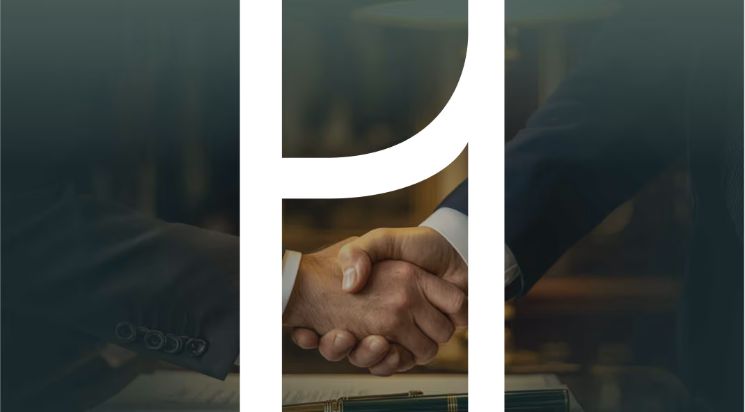 Two people in suits shaking hands over a desk with a pen and document.