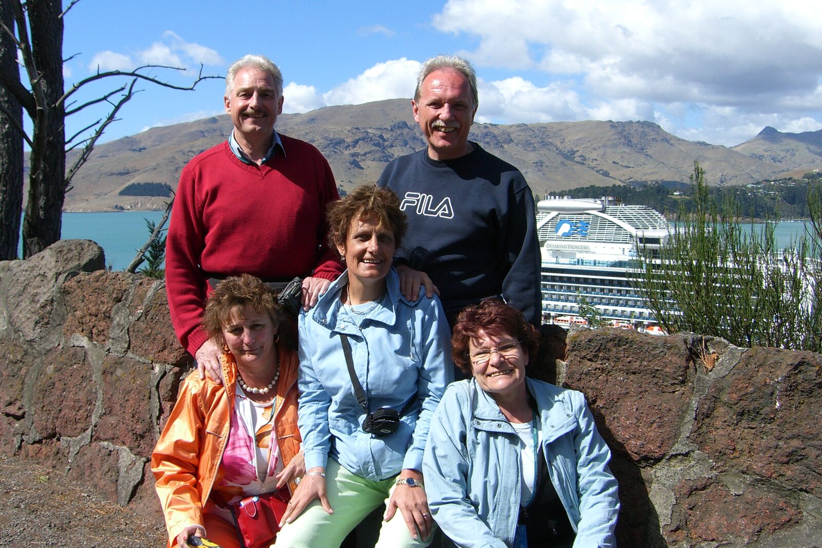 Group of five adults posing outdoors near a stone wall with a cruise ship and mountains in the background.