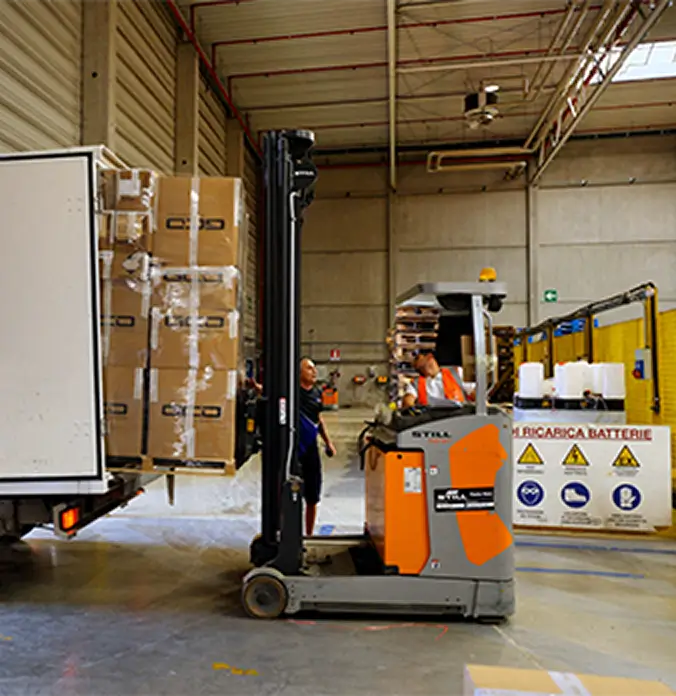 Forklift loading stacked cardboard boxes onto a truck in a warehouse with two workers nearby.