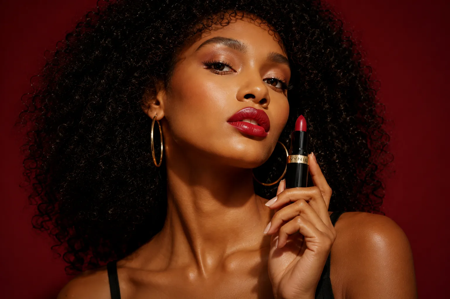 Woman with curly hair wears red lipstick and gold hoop earrings holding red lipstick tube near lips.