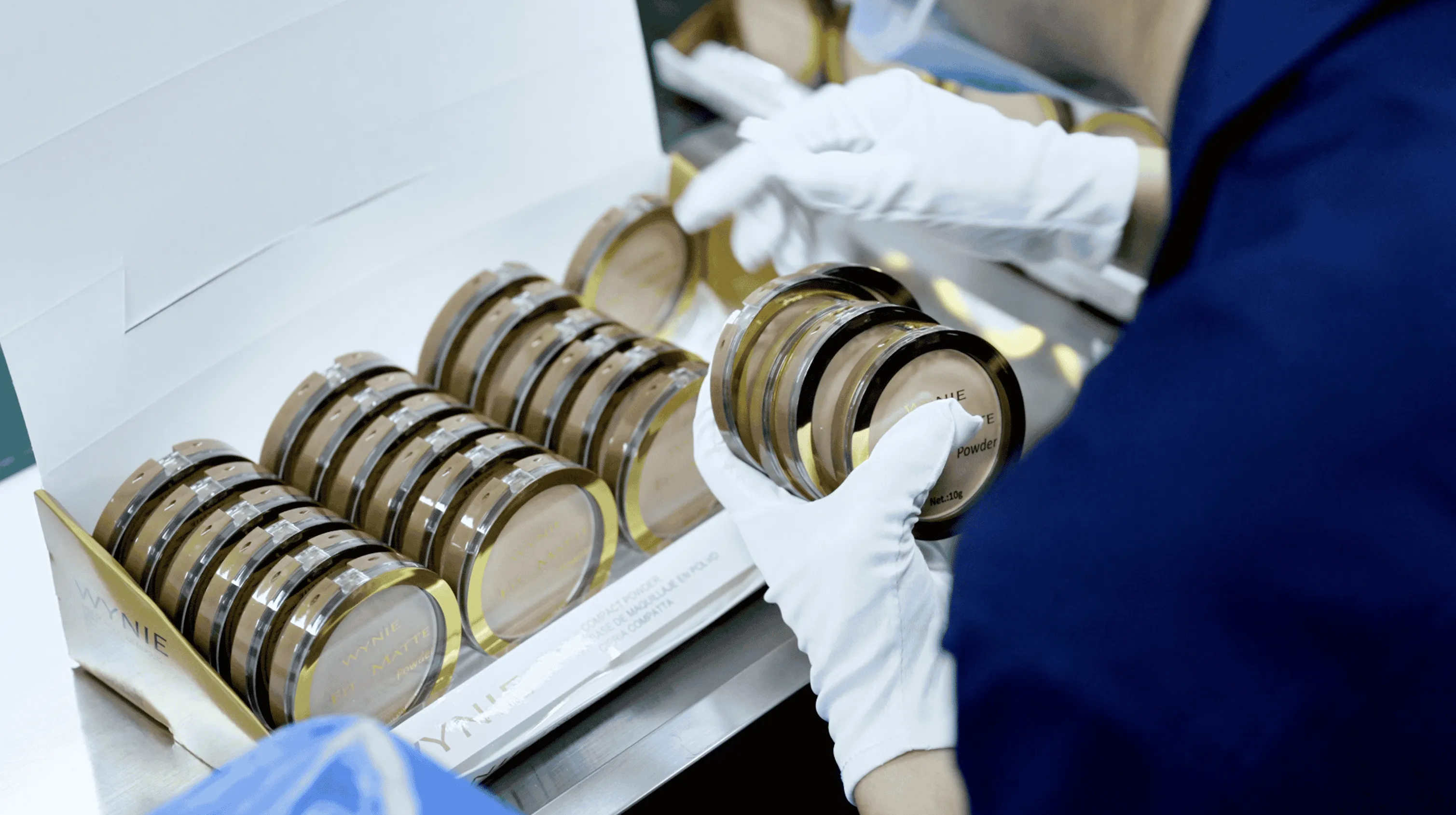 Person wearing white gloves holding compact makeup powders next to a box full of similar powders.