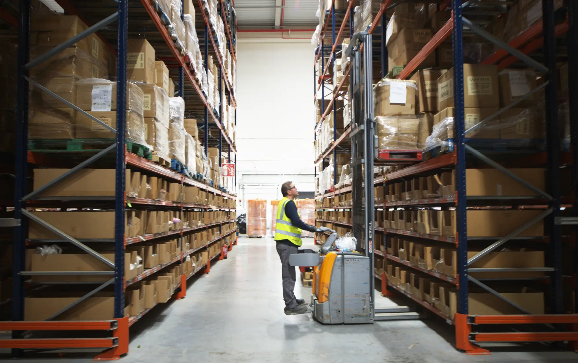 Warehouse worker with a yellow safety vest operating a forklift between tall shelves of stacked boxes.