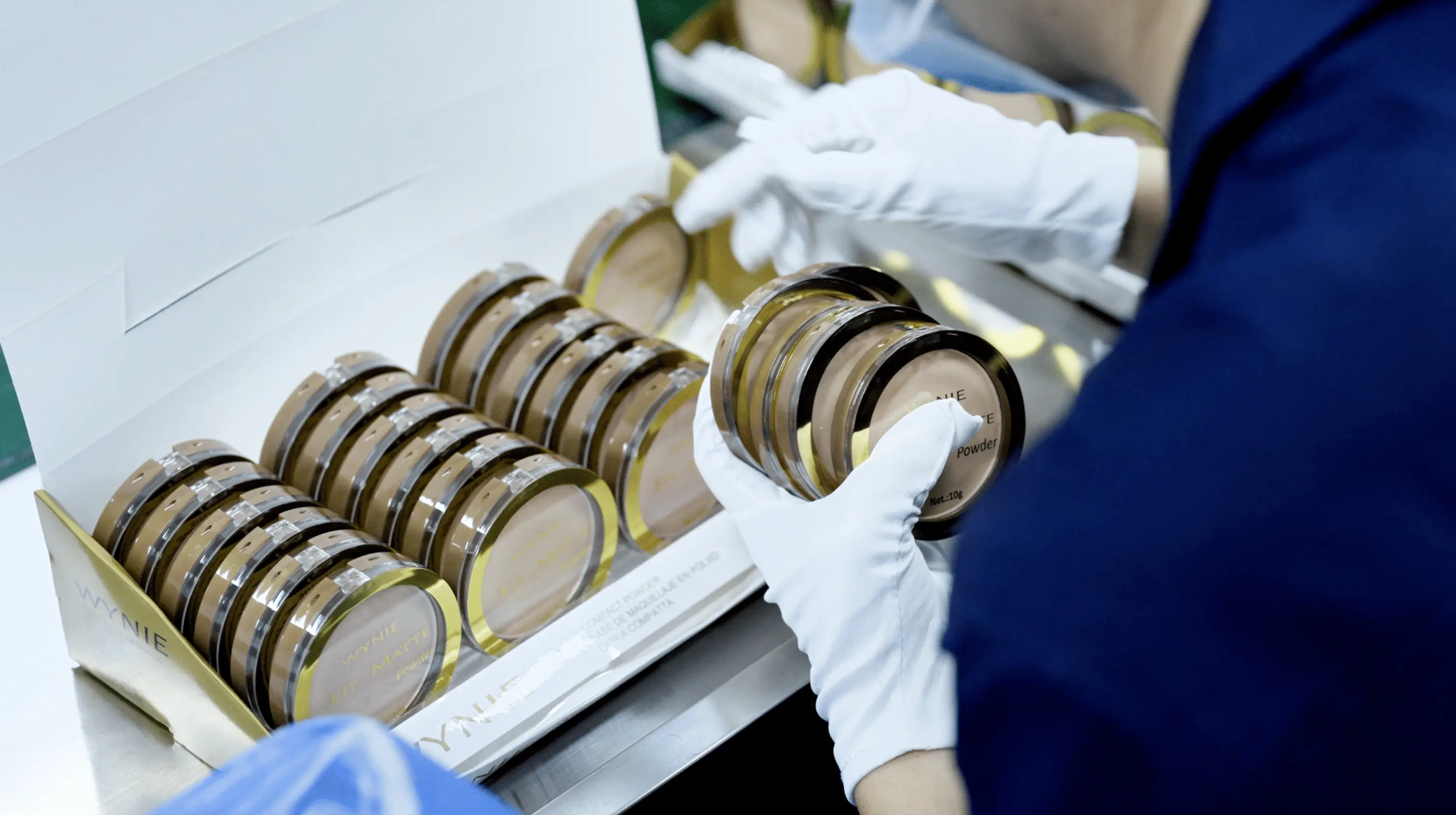 Person with white gloves holding cosmetic powder compacts from a box of neatly arranged products.