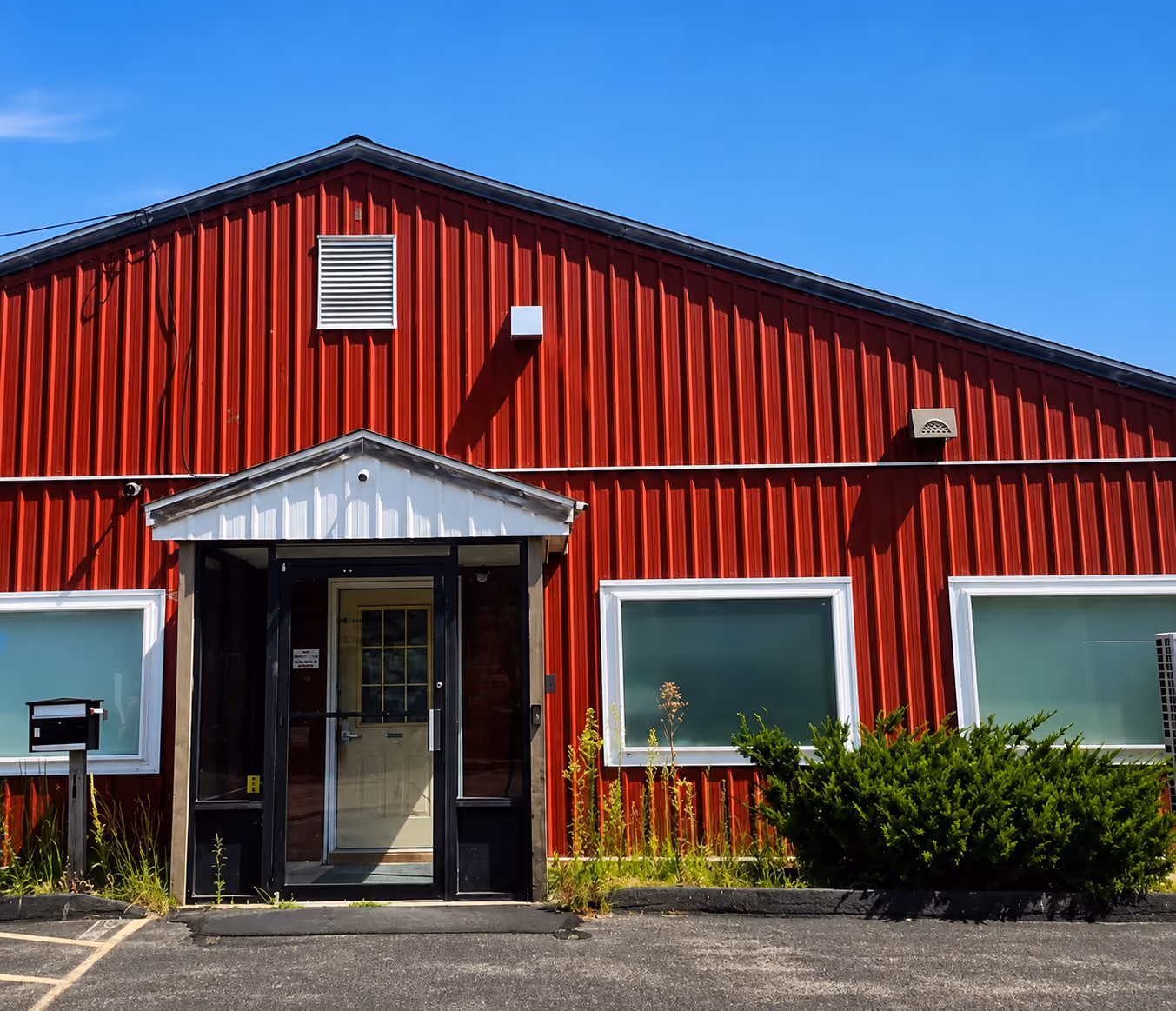 Red building with white door, two frosted windows, a mailbox, and green shrubs in front under blue sky.