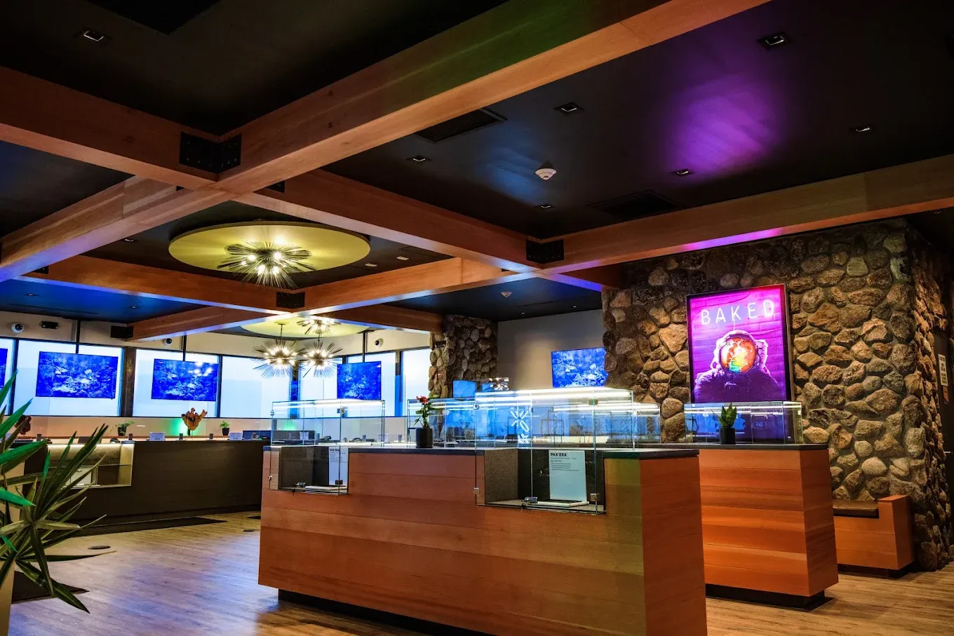 Interior of Nova Farms Framingham cannabis store, featuring wooden crossbeams, stone accent walls with a glowing "BAKED" sign, glass display cases on wooden counters, and blue display screens in the background.