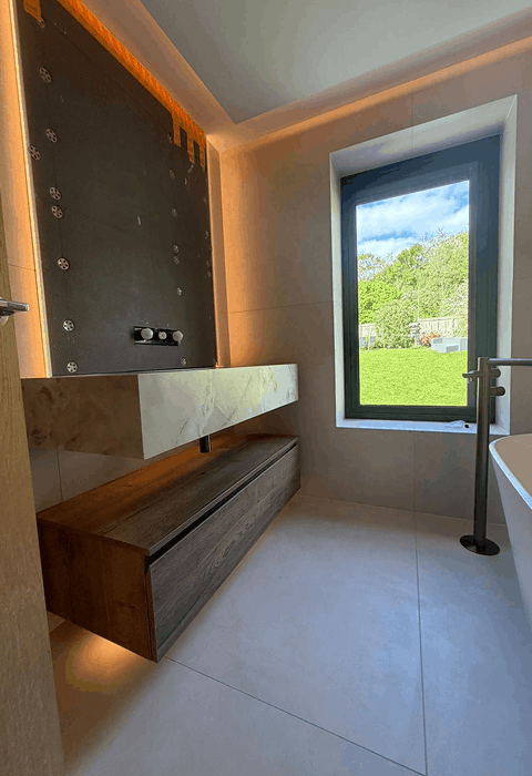 Modern bathroom at night with a large white freestanding bathtub on a dark rug, a glass shower enclosure, double sink vanity, lit fireplace, and a woman in a robe standing near tall windows with curtains.