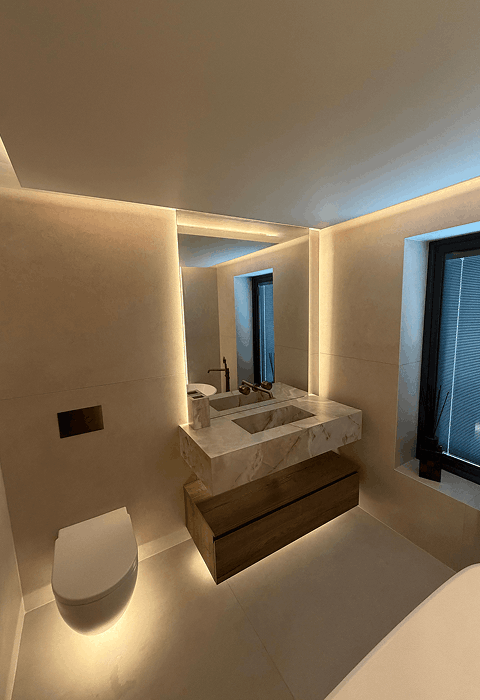 Modern bathroom at night with a large white freestanding bathtub on a dark rug, a glass shower enclosure, double sink vanity, lit fireplace, and a woman in a robe standing near tall windows with curtains.