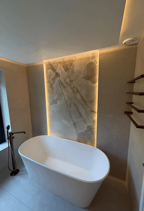Modern bathroom at night with a large white freestanding bathtub on a dark rug, a glass shower enclosure, double sink vanity, lit fireplace, and a woman in a robe standing near tall windows with curtains.