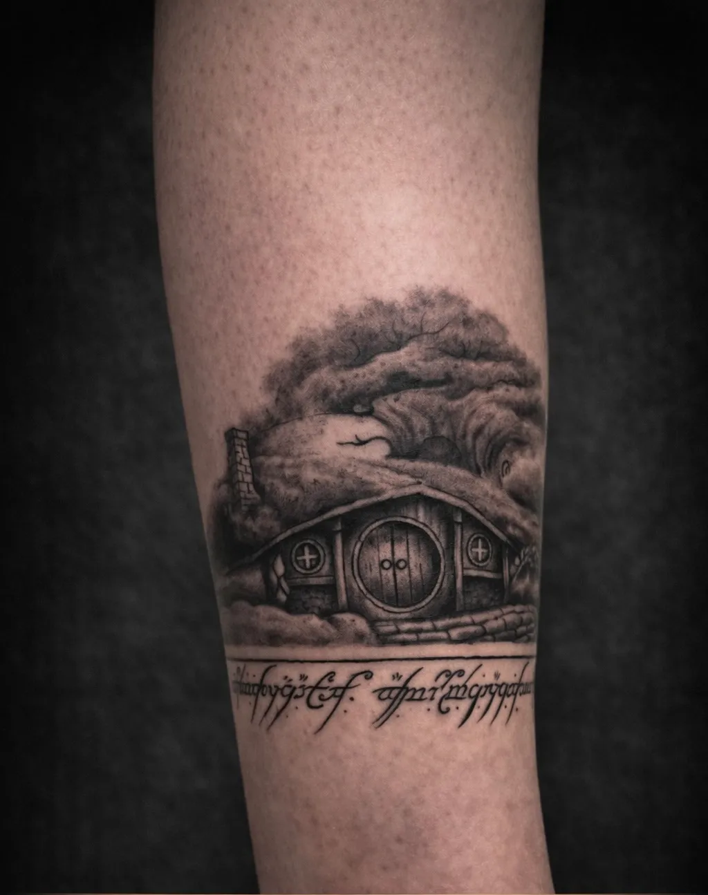 Tattoo on a forearm showing a detailed black and grey depiction of a Hobbit-style house with a round door, windows, a stone chimney, and a large gnarled tree behind it, with Elvish script below the house.