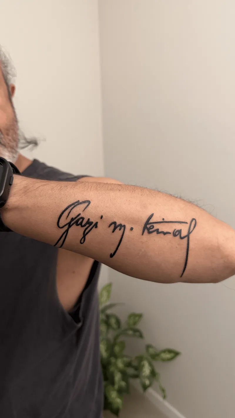 Arm mit einem Tattoo der Unterschrift "Gazi M. Kemal" in schwarzer Tinte auf der Innenseite des Unterarms, eine Person trägt ein schwarzes Armband und ein dunkles ärmelloses Shirt.