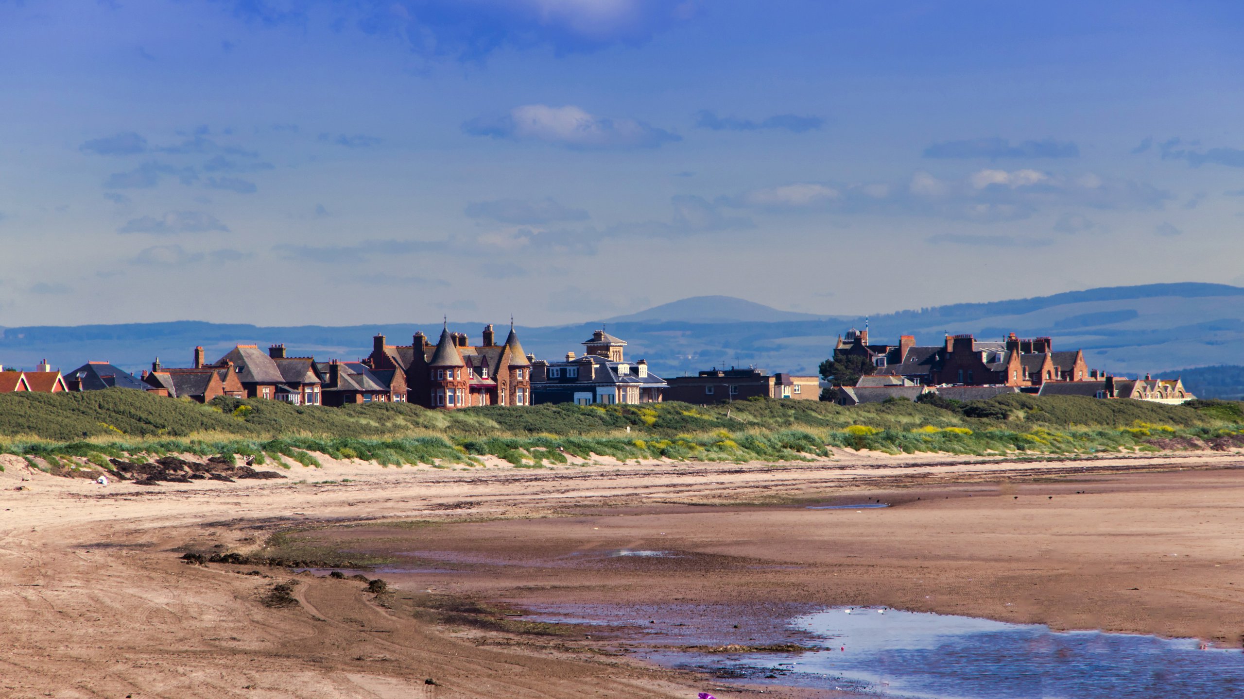 Coastal town with sandy beach, green dunes, and brick houses under a blue sky with clouds.