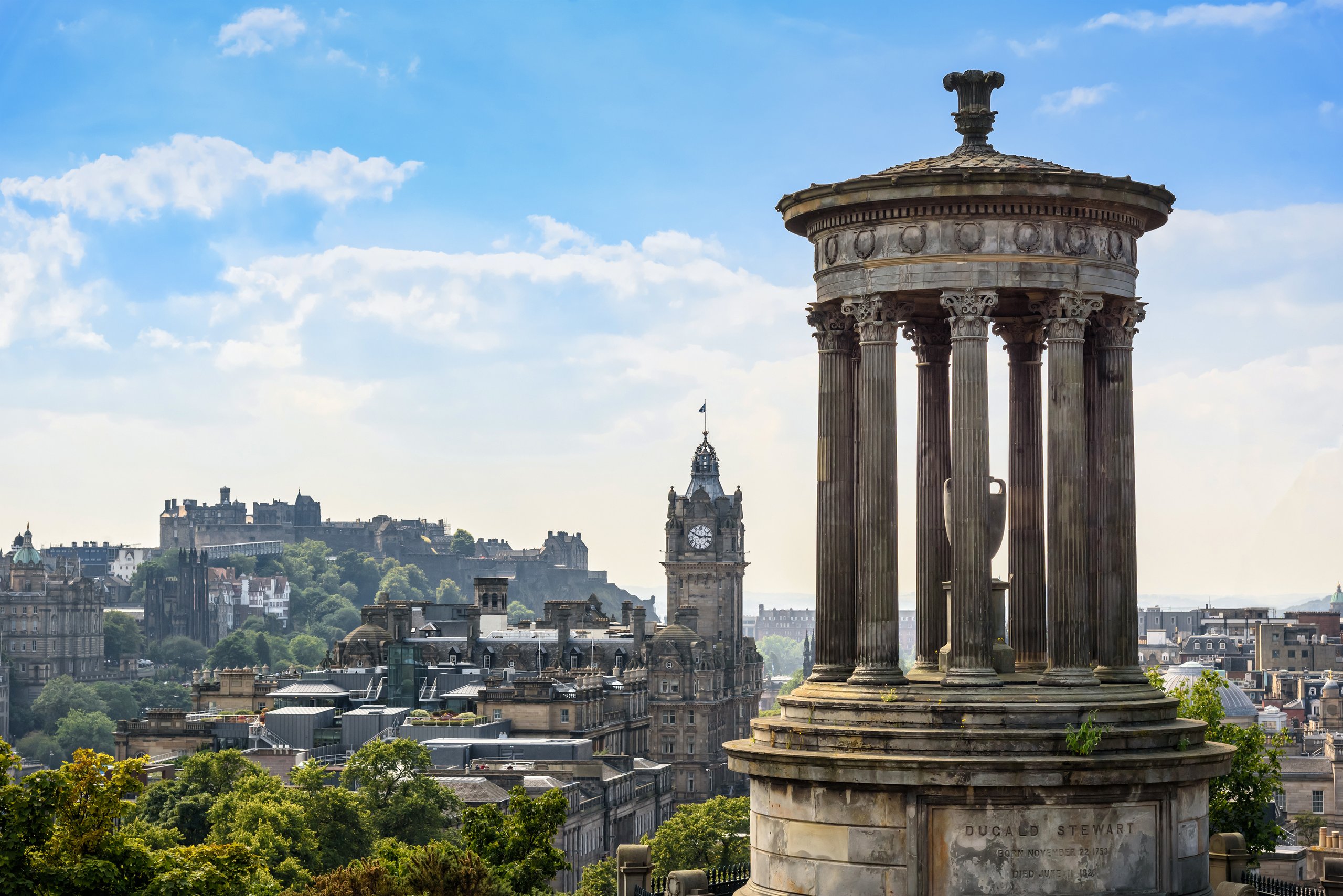 View of Calton Hill monument with Edinburgh cityscape and castle in the background under blue sky.