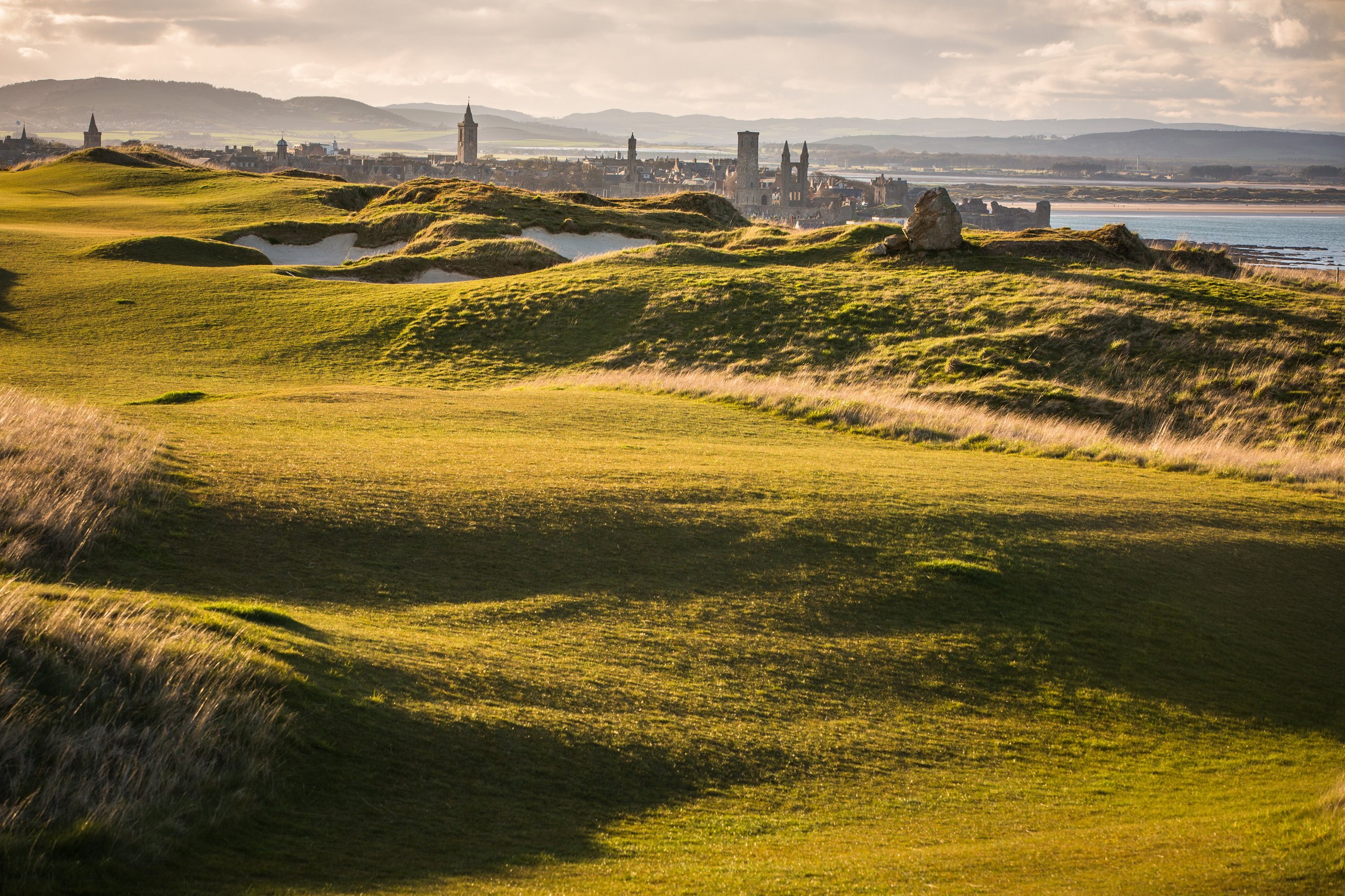 Rolling green golf course with sand bunkers overlooking a distant town and coastline under a cloudy sky.
