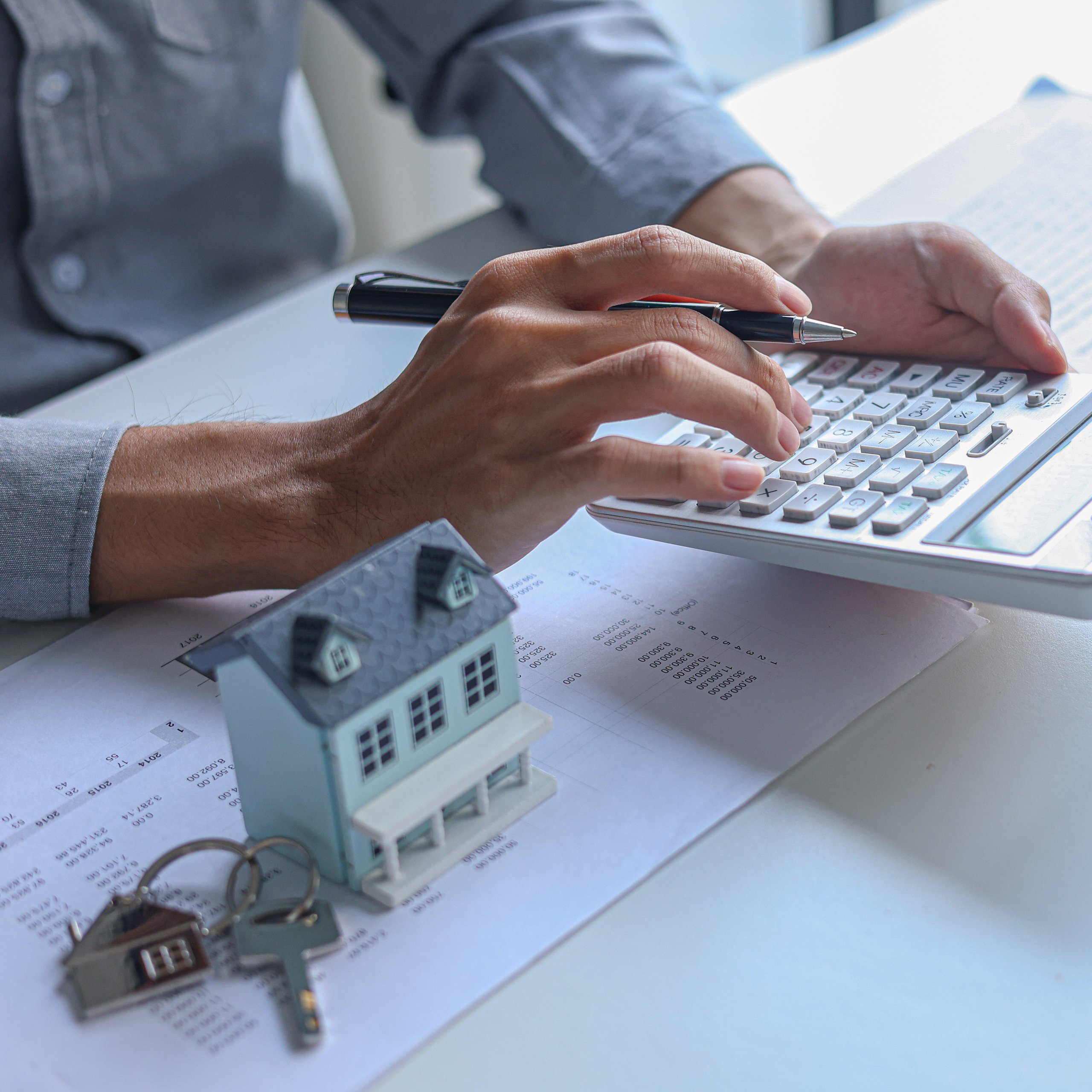 Person calculating finances with a pen and calculator near a house model and keys on papers.