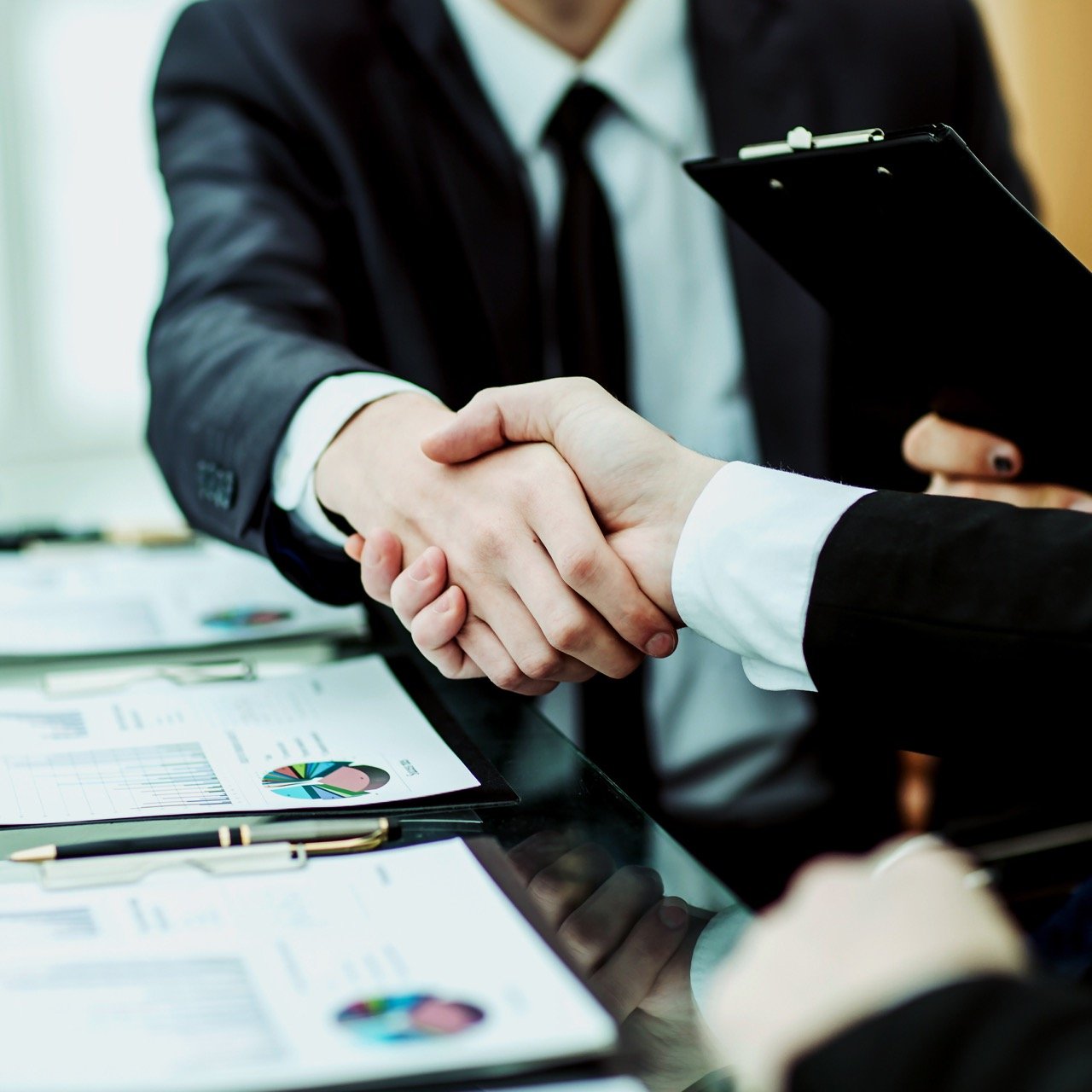 Two businesspeople in suits shaking hands over a glass table with charts and documents.