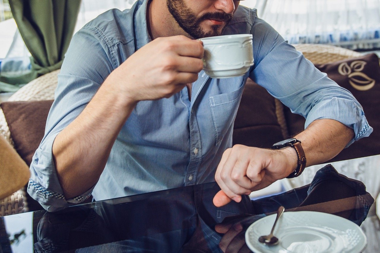 Man in blue shirt holding a cup and checking his wristwatch while sitting at a table with a plate and spoon.