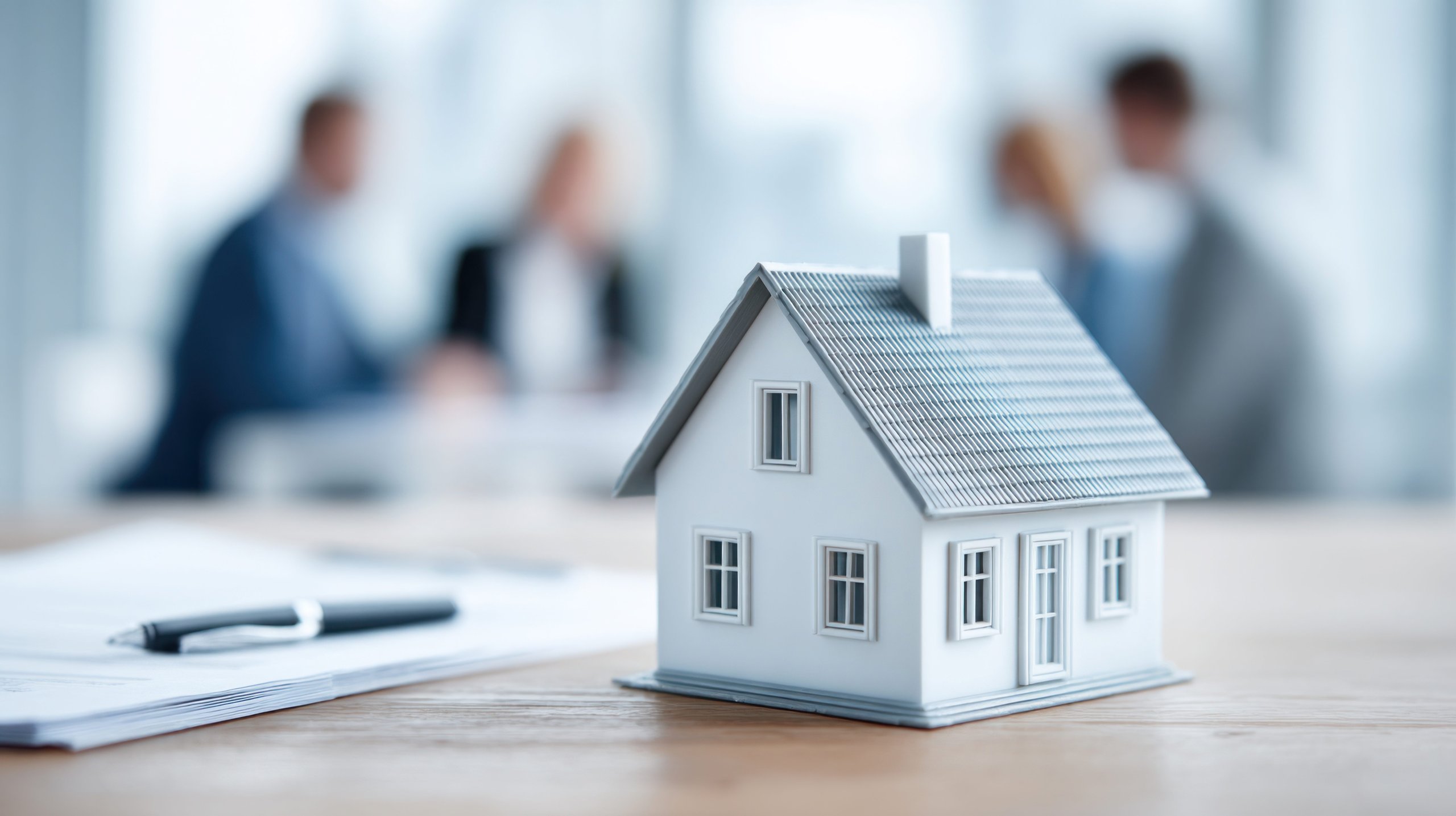 Small white model house on desk with pen, papers, and blurred people in background.